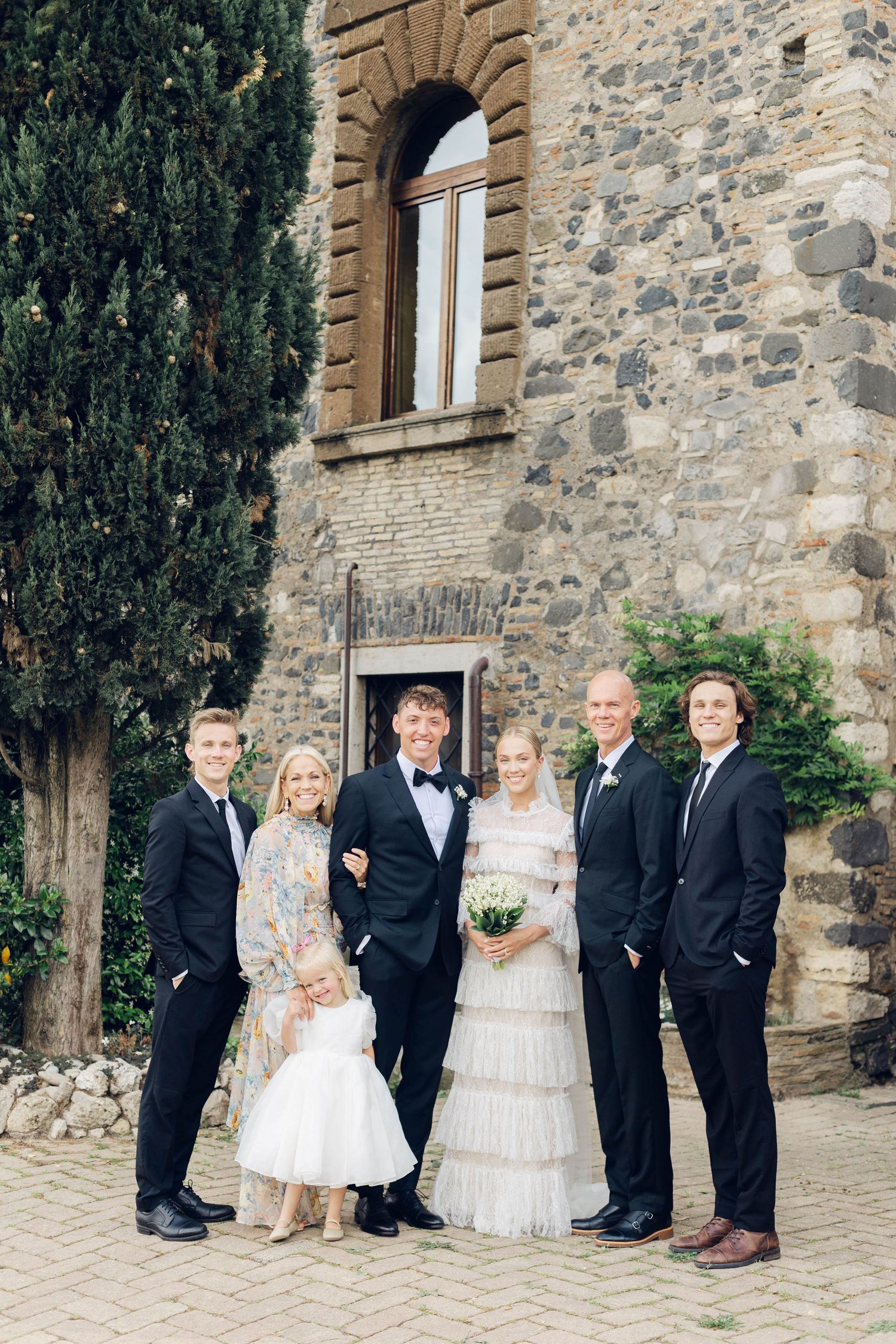 Wedding party poses together outside stone villa with cypress trees.