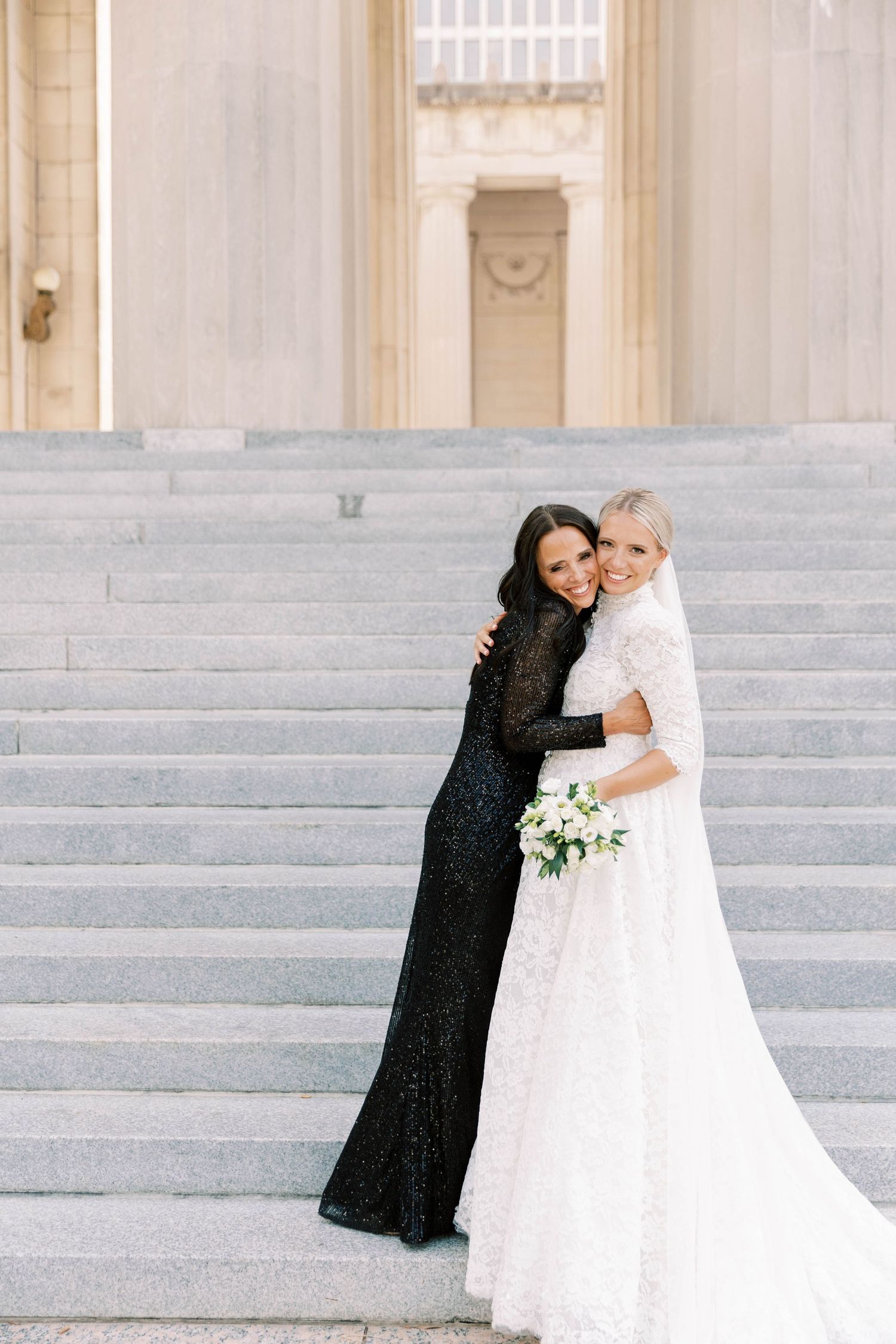 Bride in white gown and friend in black dress embrace on grand marble steps.