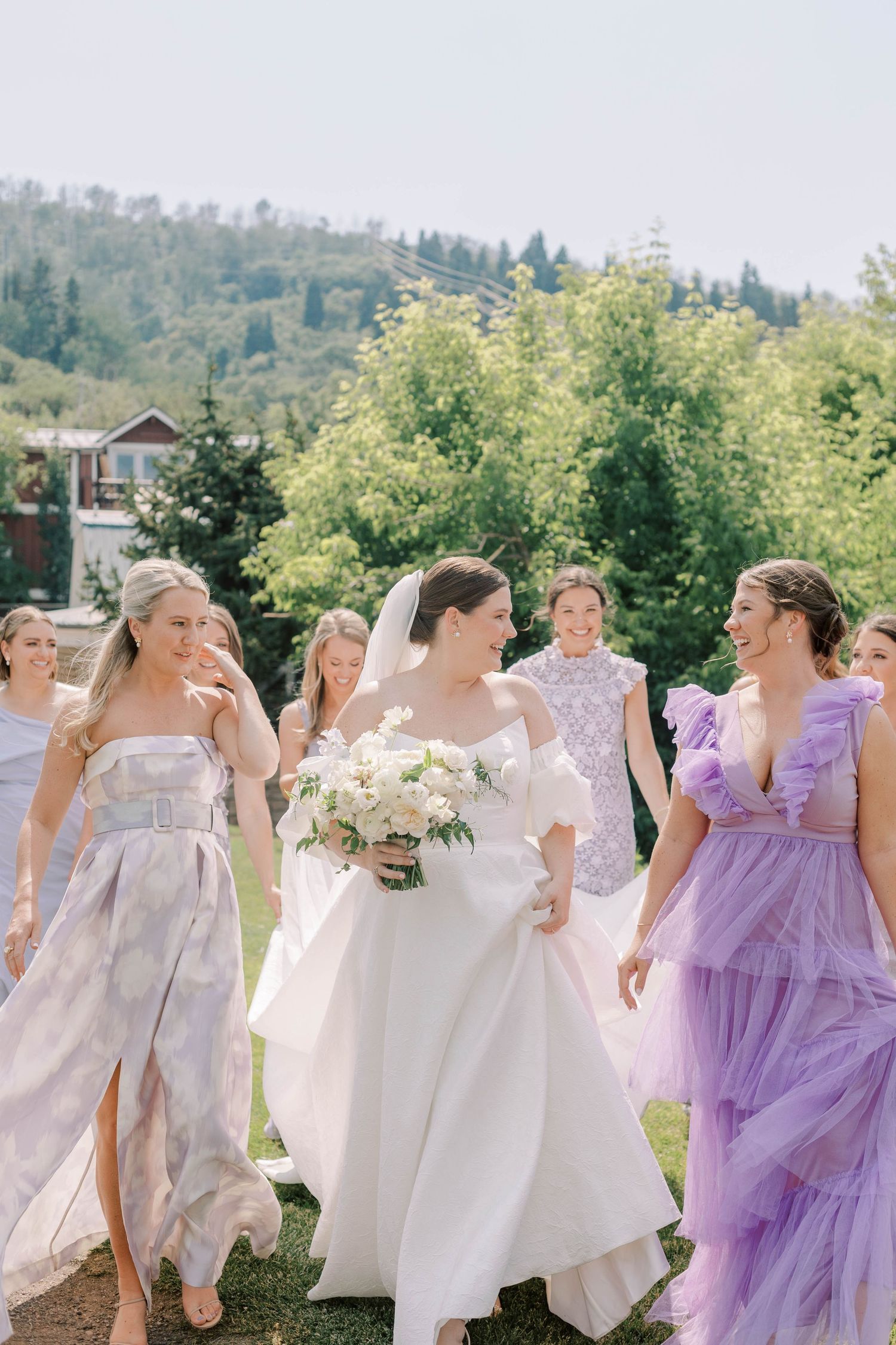 Bridesmaids in pastel dresses walk with bride carrying white bouquet in outdoor setting.