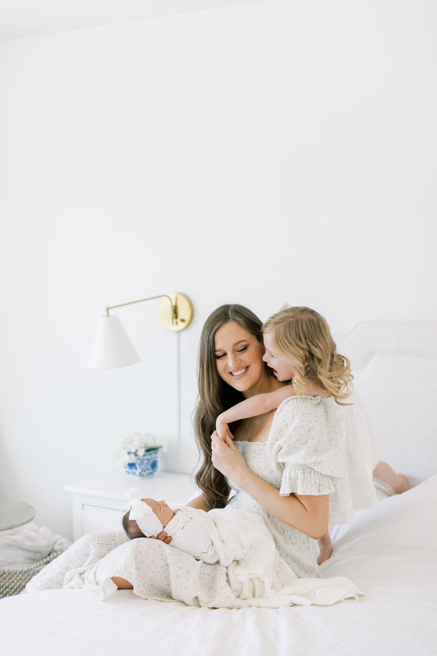 Mother and daughter in white dresses sharing a tender moment on a bed with a gold lamp nearby.