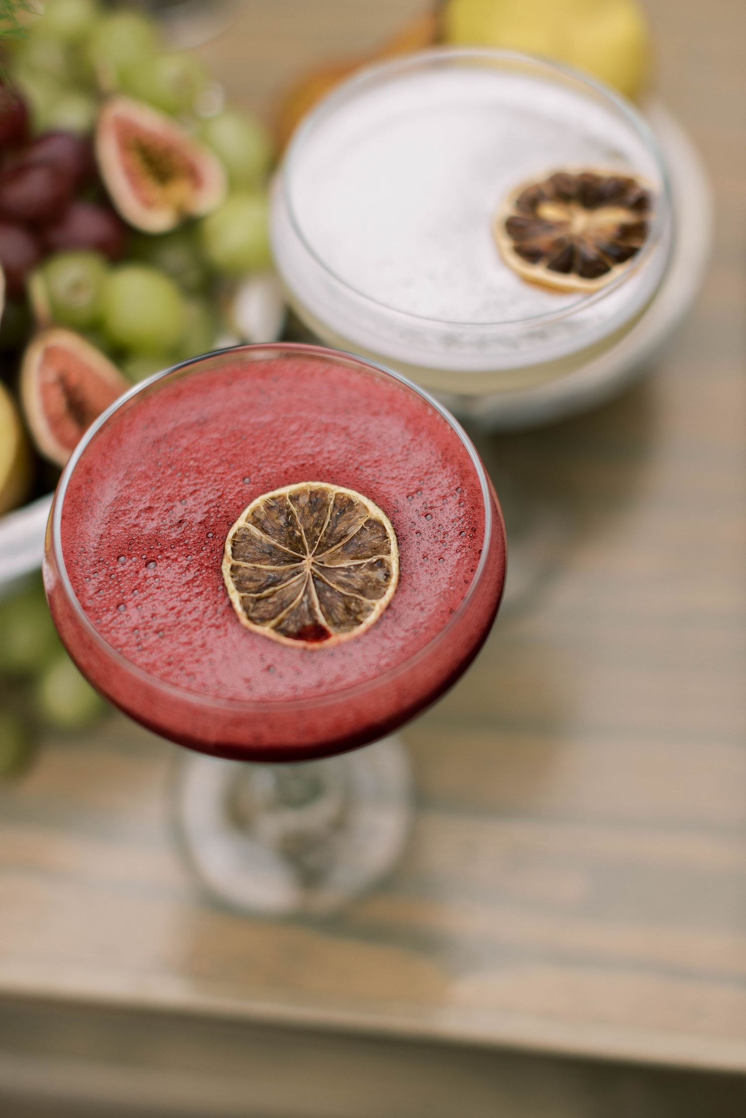 Two cocktails garnished with dried citrus slices, one pink and one white, surrounded by fresh fruit.