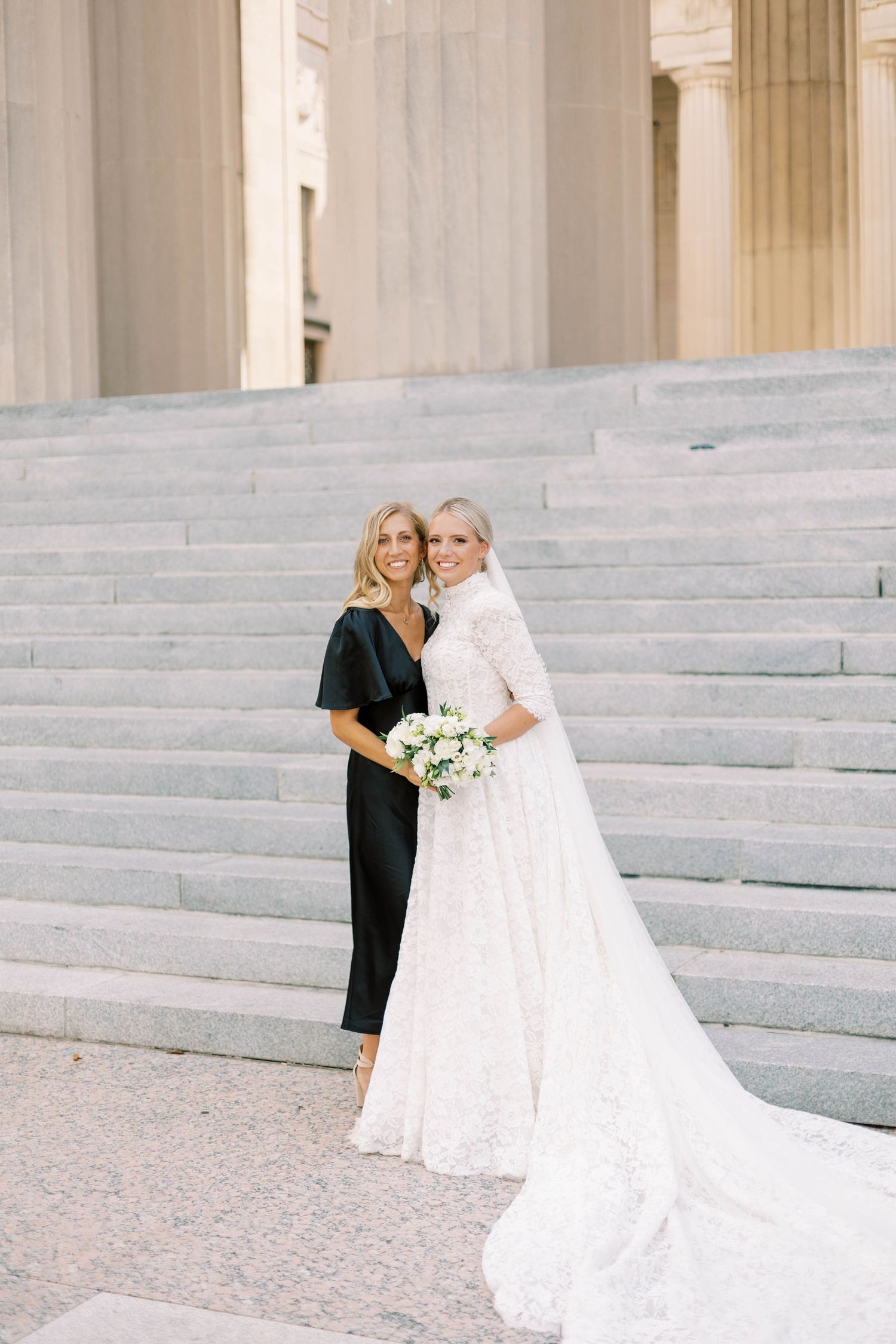 Bride in white lace gown poses with attendant in black dress on grand stone steps.