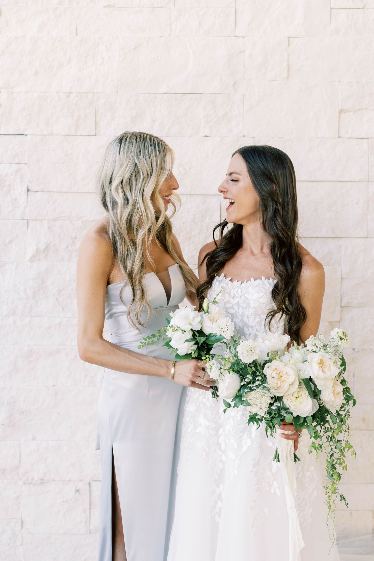Bride in white lace gown with bridesmaid in silvery dress sharing joyful moment while holding elegant floral bouquet.