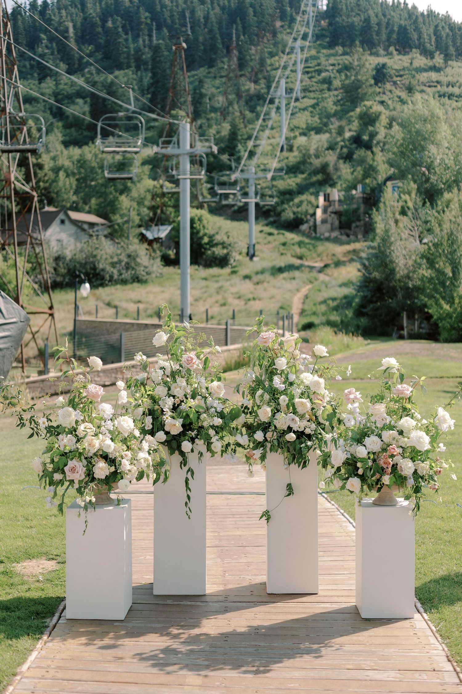 Four white pedestals adorned with lush floral arrangements in front of a ski lift on a mountain slope.