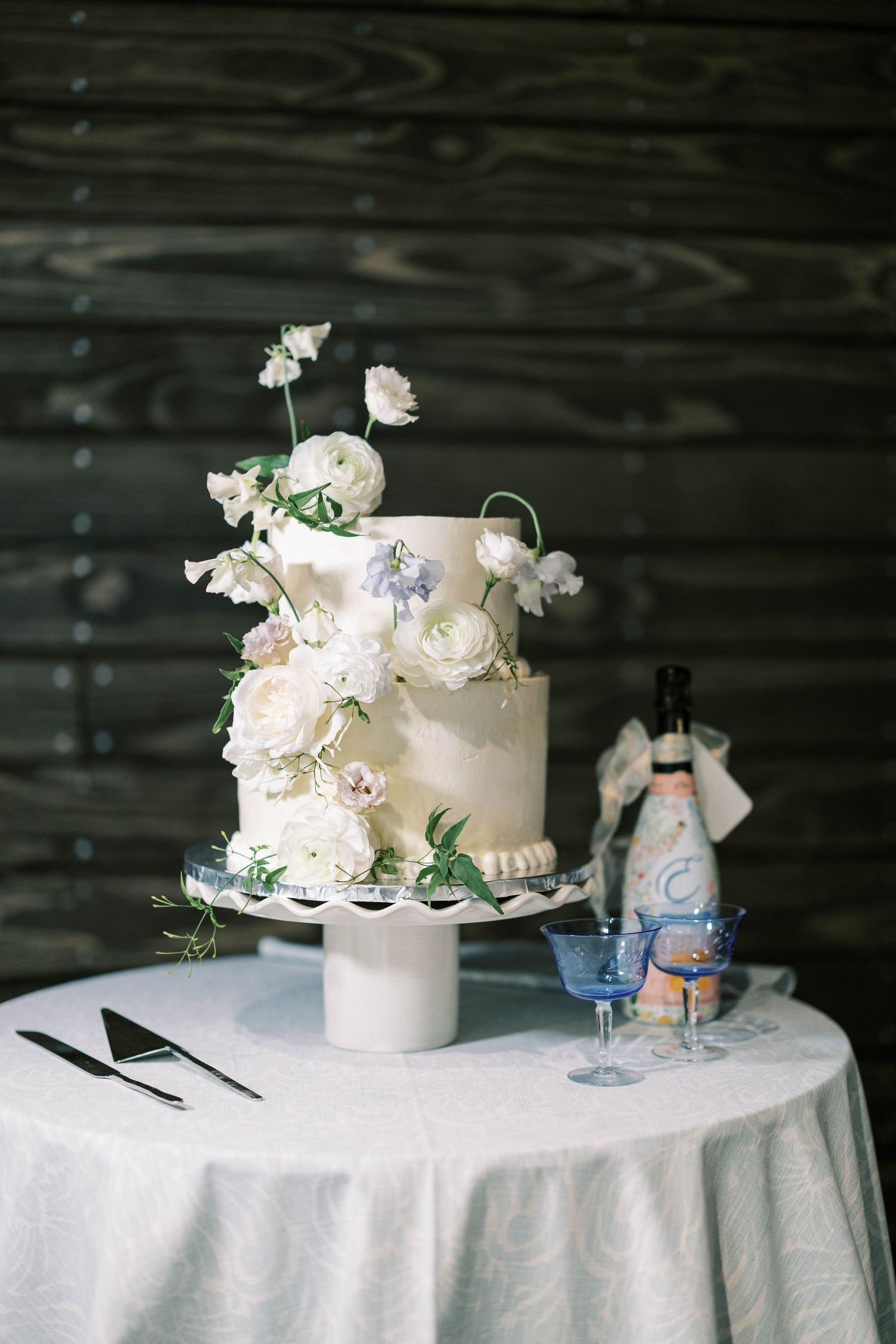 Elegant white wedding cake decorated with fresh flowers and greenery on round table.