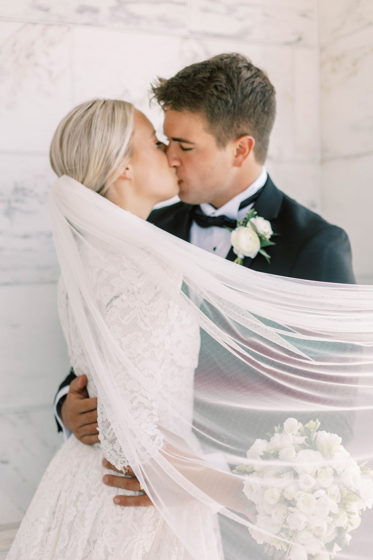 Romantic wedding kiss framed by flowing veil against marble wall.