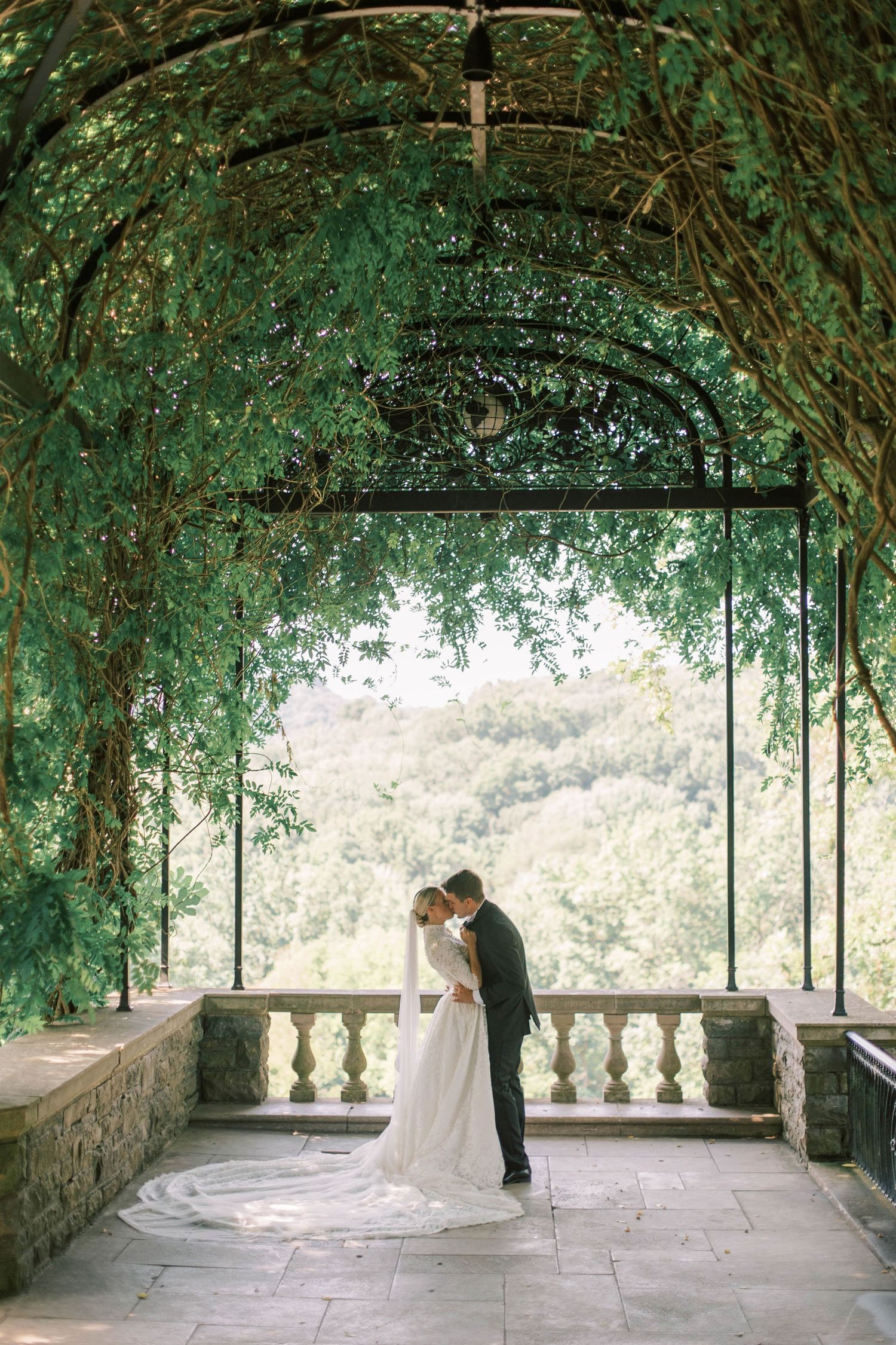 Wedding couple embraces under vine-covered stone arbor overlooking scenic valley of Cheekwood Estate.