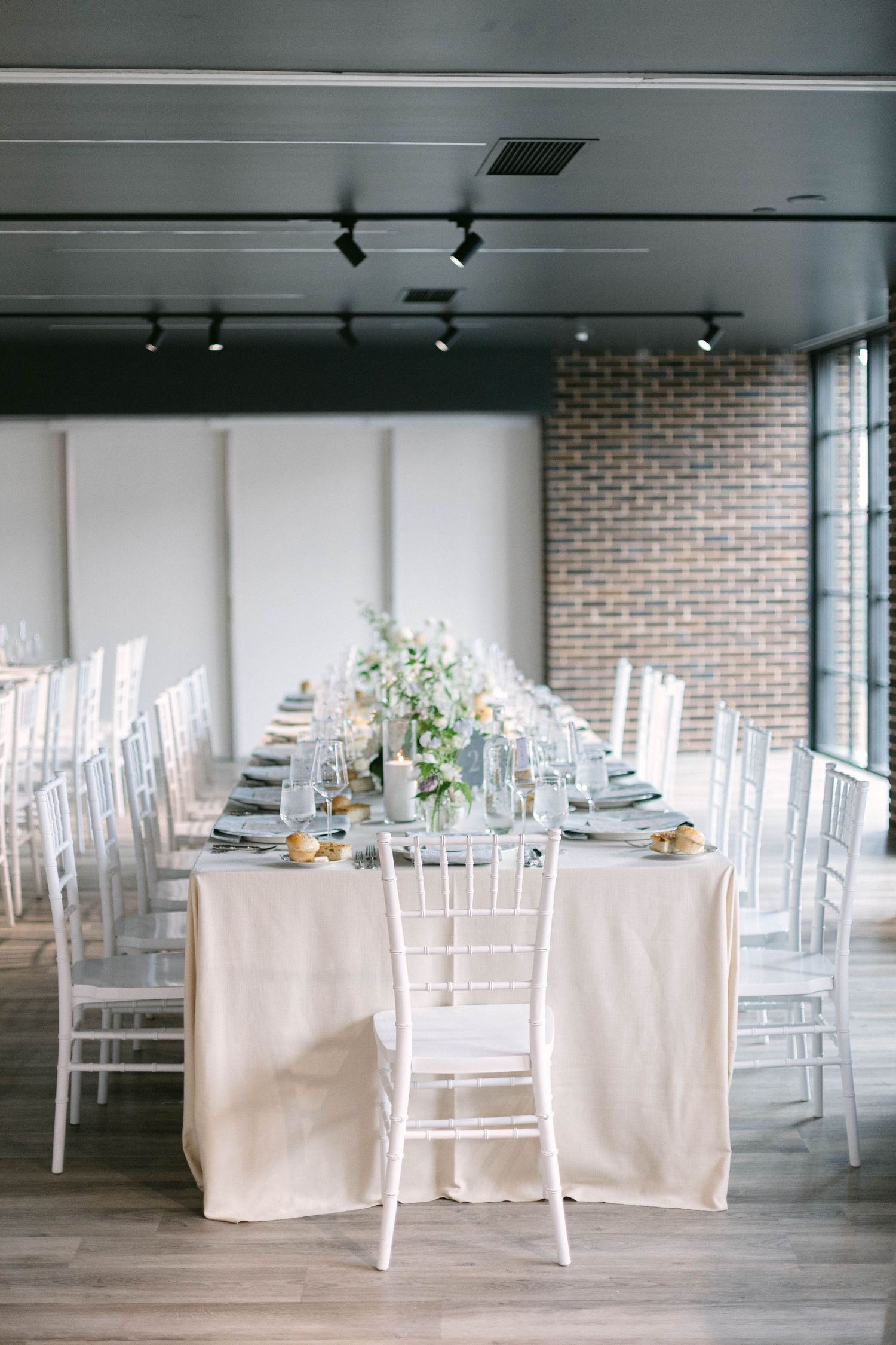 Long dining table with white chiavari chairs and floral centerpieces in an industrial-style venue.