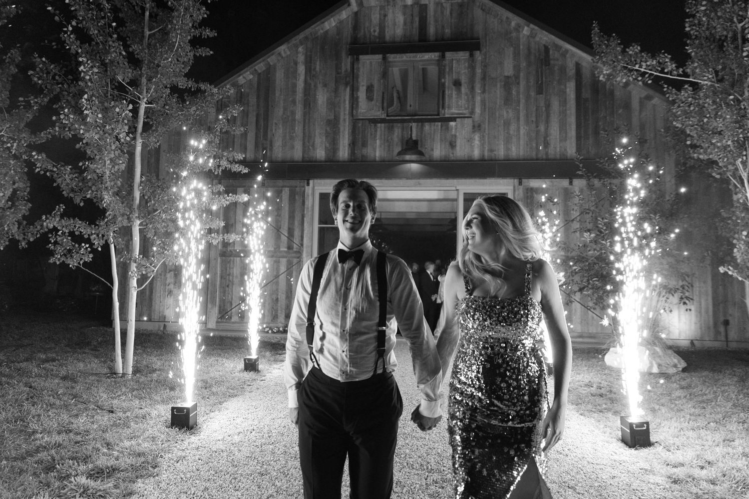 Black and white photo of couple walking past sparkler fountains in front of rustic barn venue.
