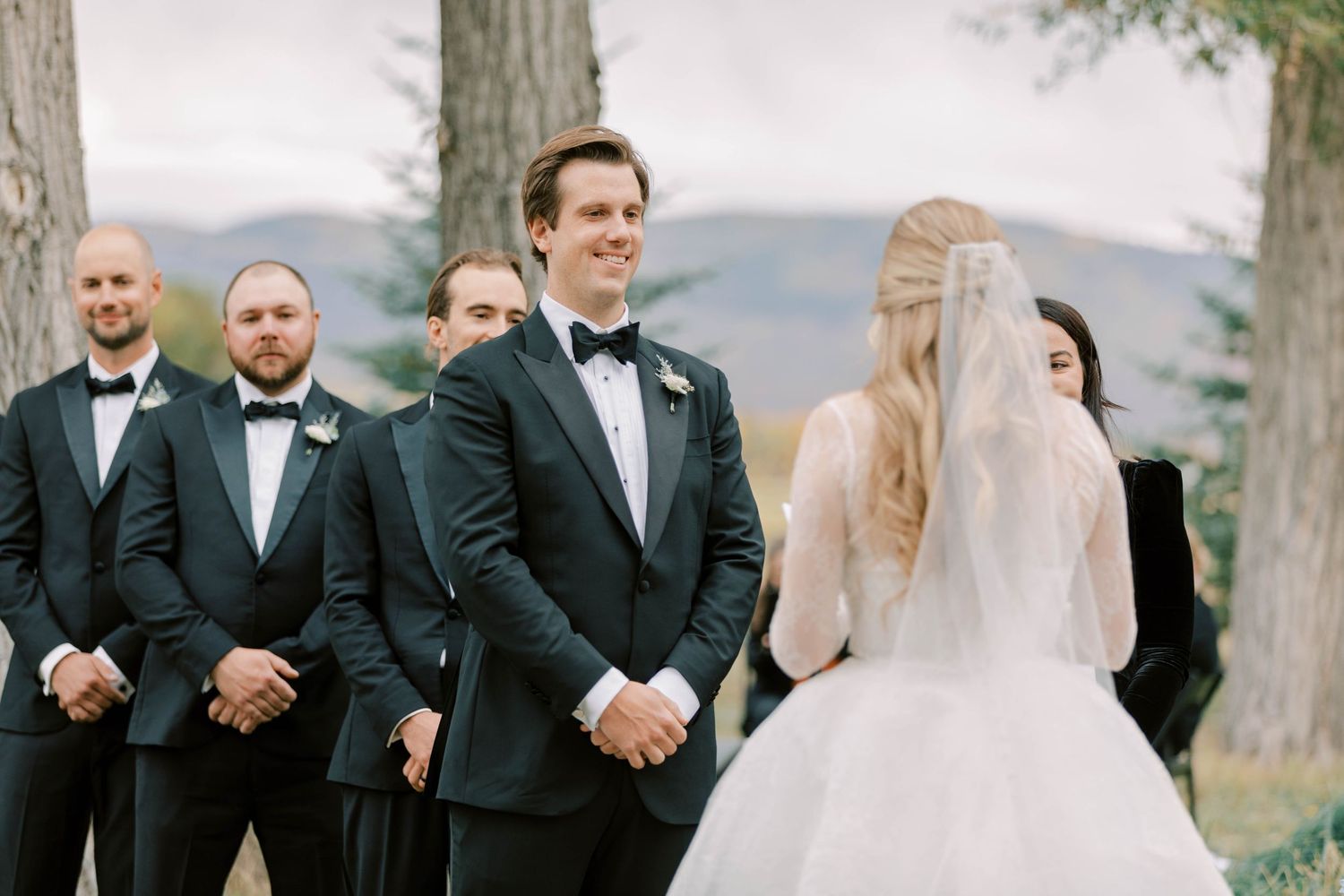 A wedding ceremony outdoors with groomsmen in black tuxedos standing near a bride in a white dress with mountains in background.