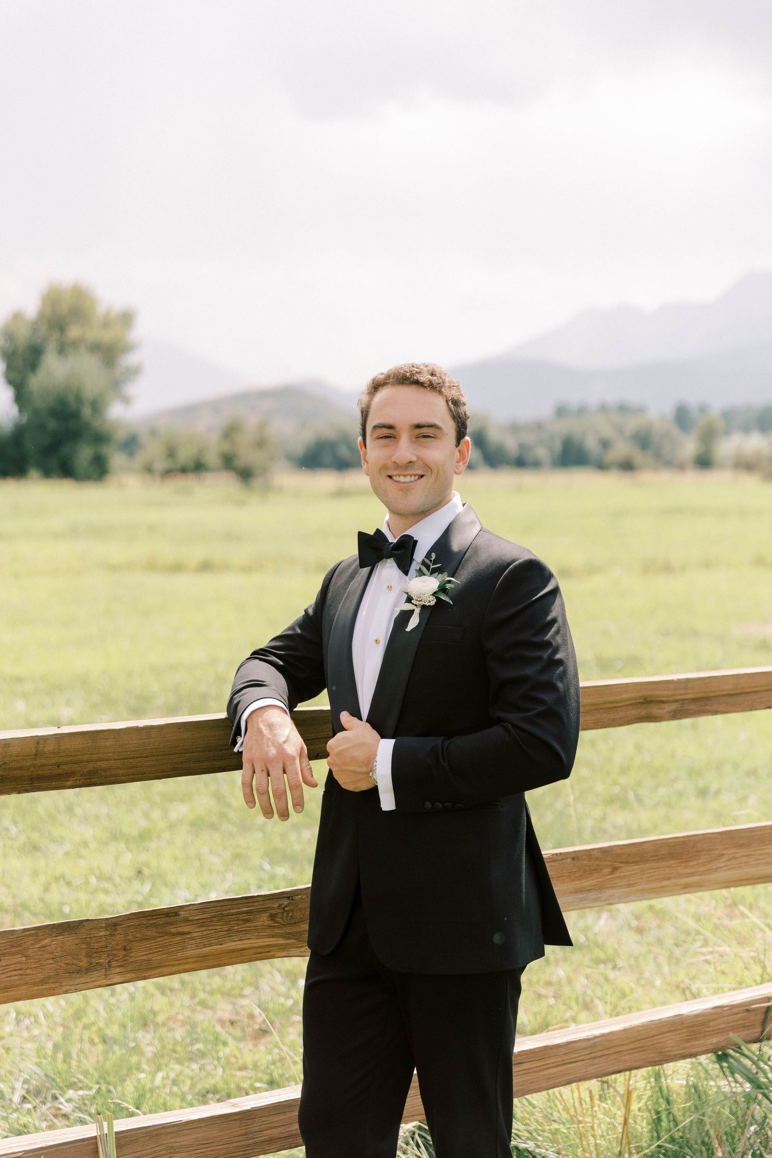 Groom in black tuxedo poses against wooden fence with mountain landscape.