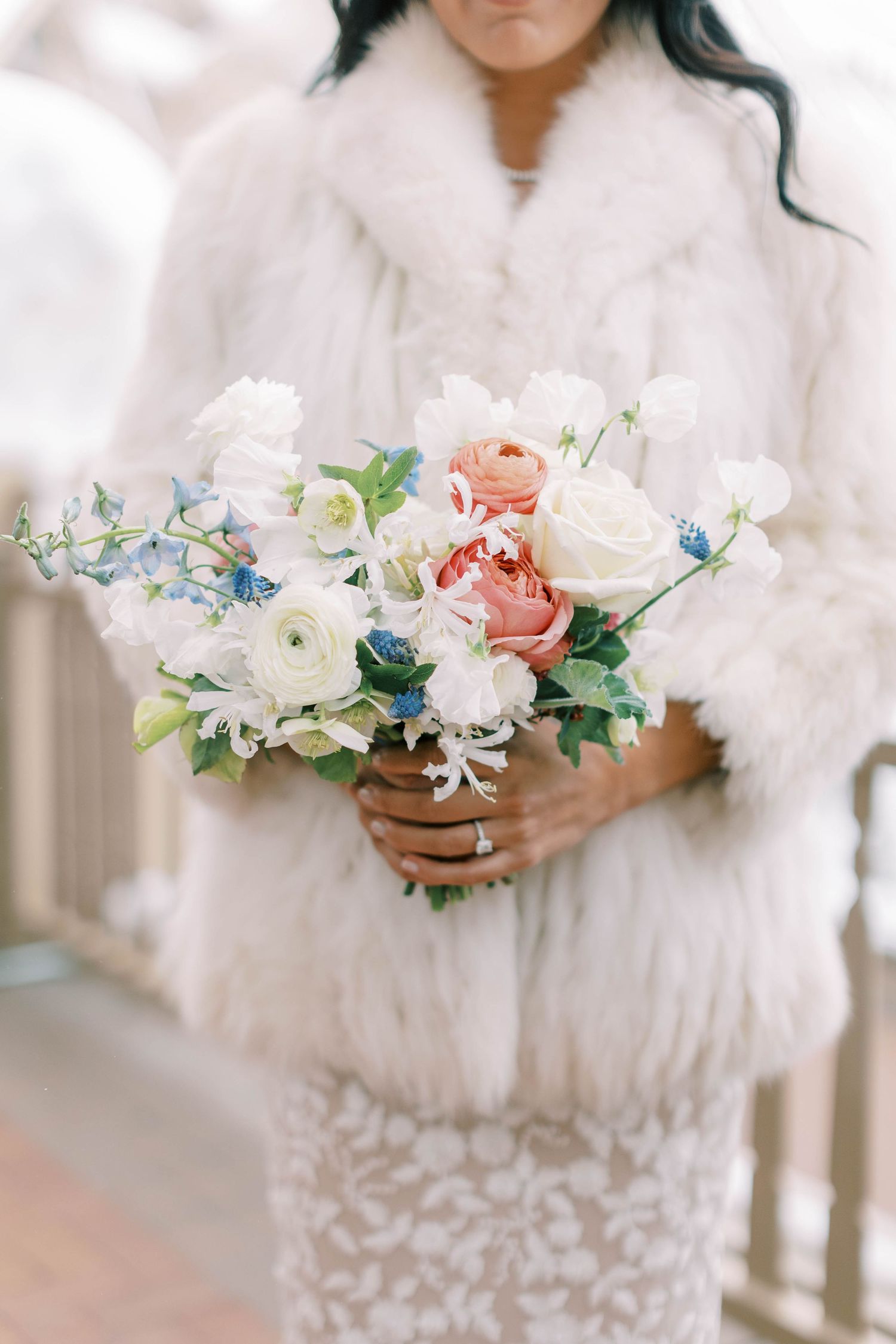 A delicate bridal bouquet featuring pink roses, white flowers and blue accents held against a white fur coat.
