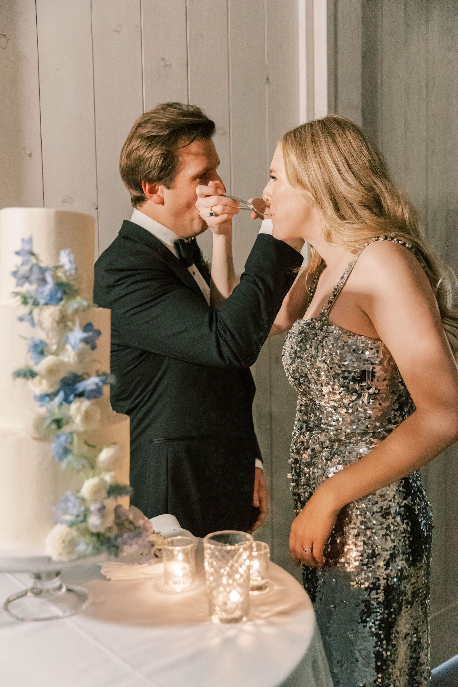 Playful wedding couple feeding each other cake beside their elegant floral-decorated wedding dessert.