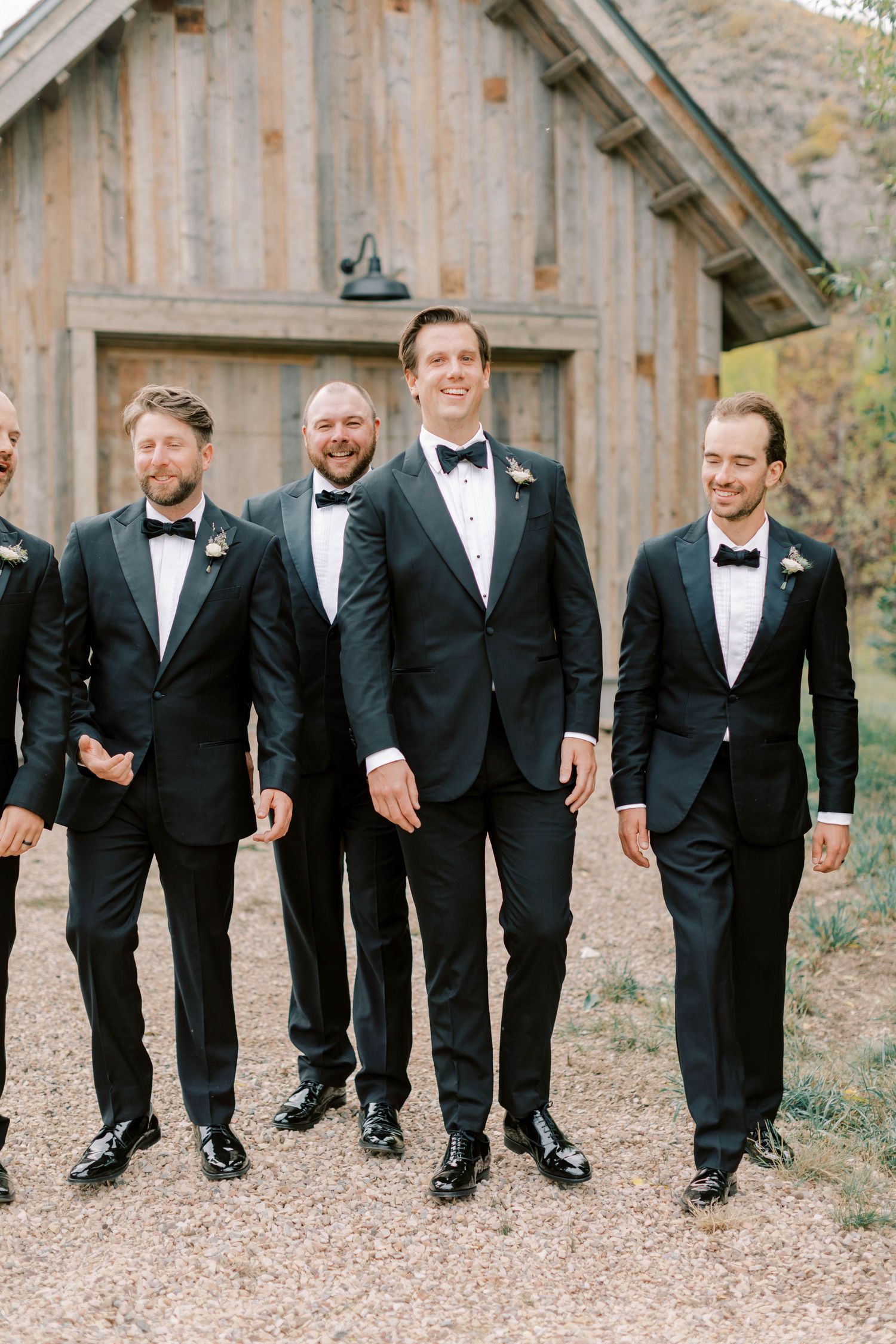 A group of groomsmen in black tuxedos and bow ties smile while walking together near a rustic barn.