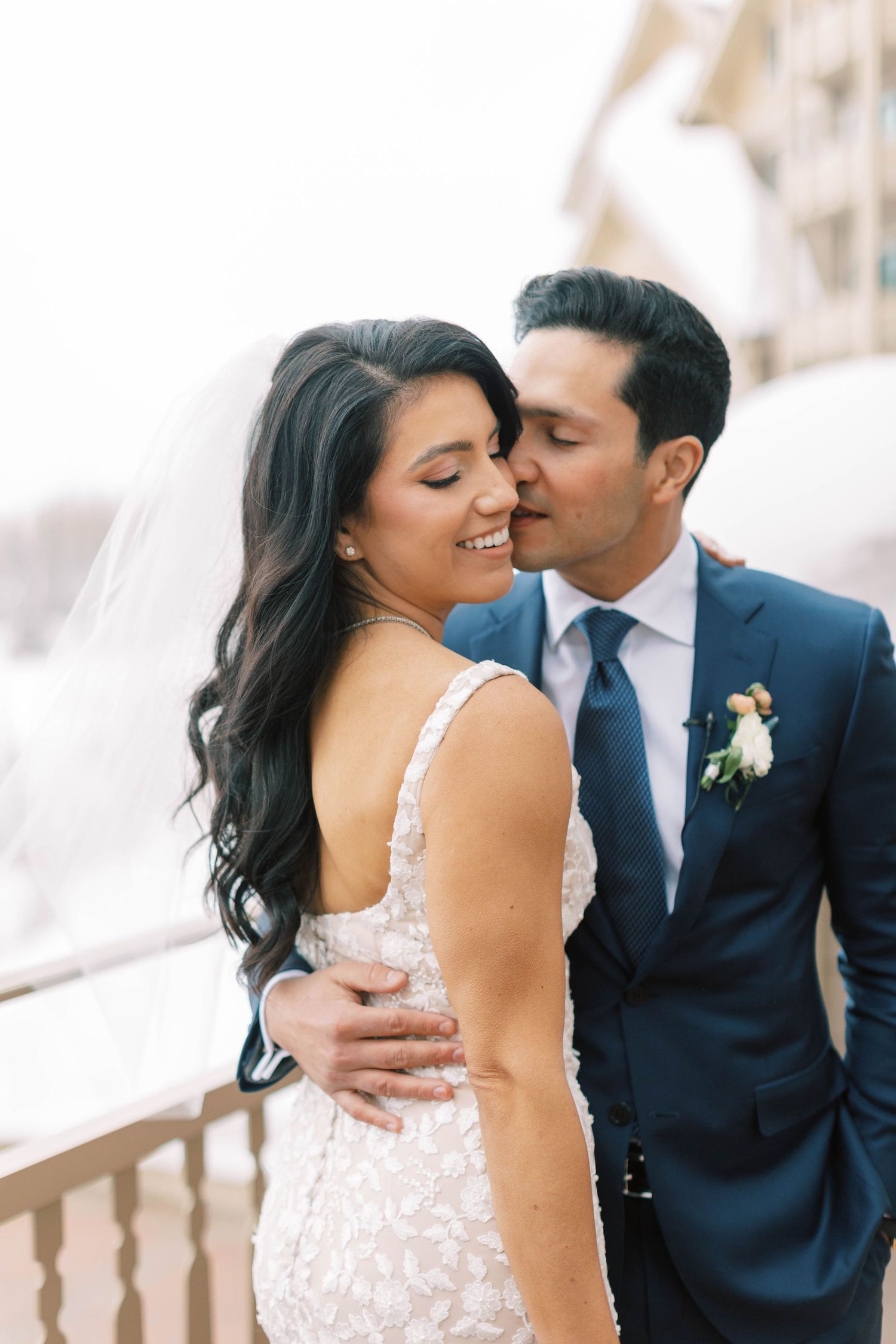 A romantic wedding moment captured as a couple in formal attire share an intimate embrace on a balcony.