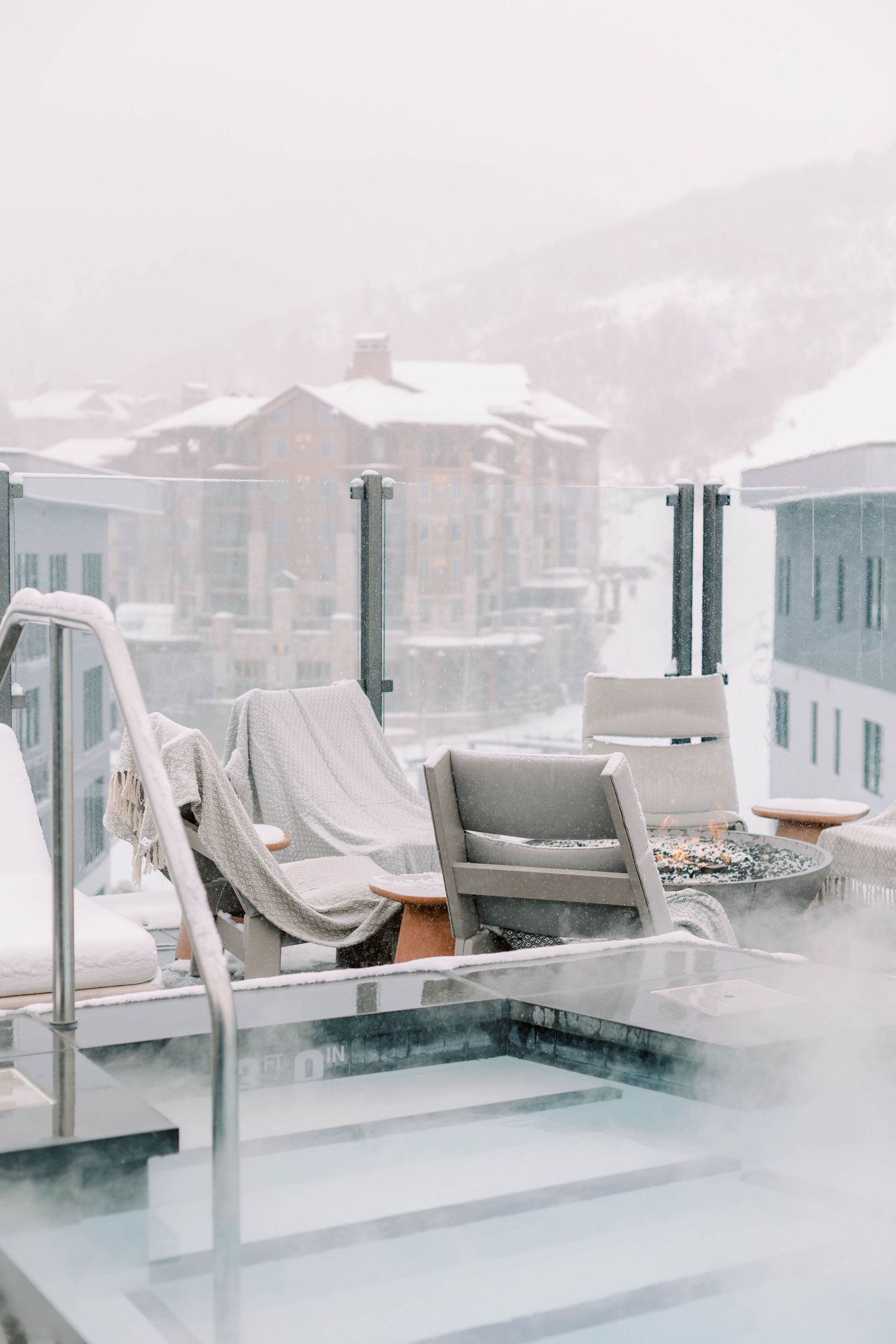 Snow-covered rooftop terrace with lounge chairs overlooking a winter mountain resort.