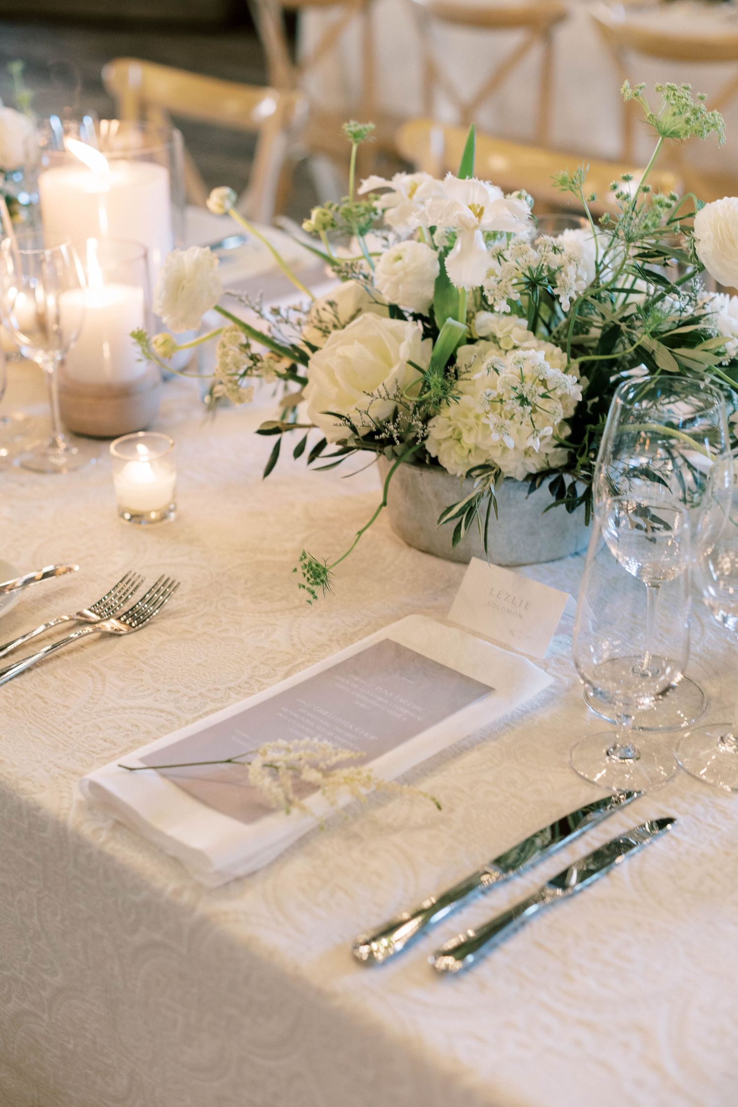 White floral centerpiece with delicate greenery on textured tablecloth with place settings.
