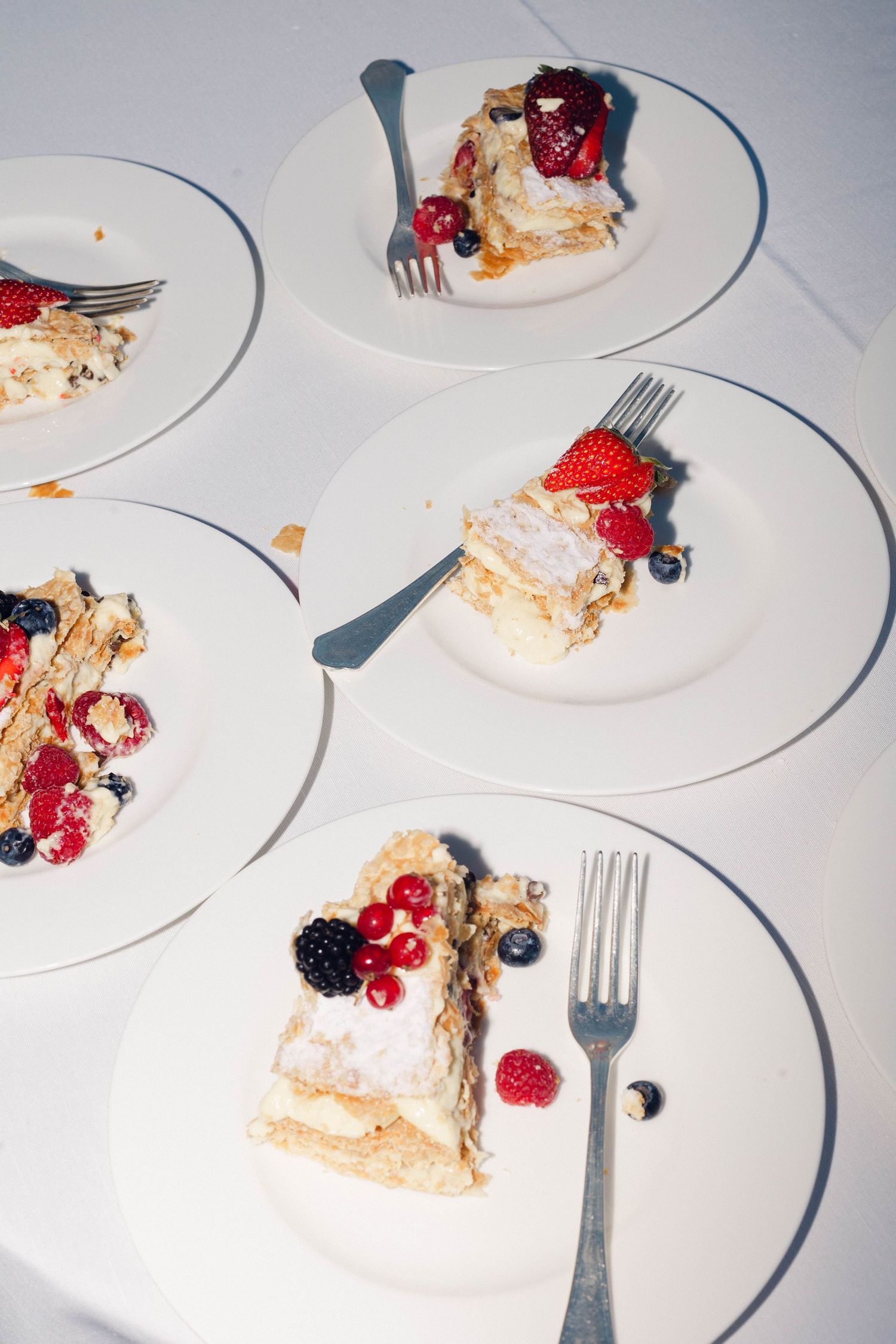 Multiple plates of layered dessert garnished with fresh berries and forks on white tablecloth.