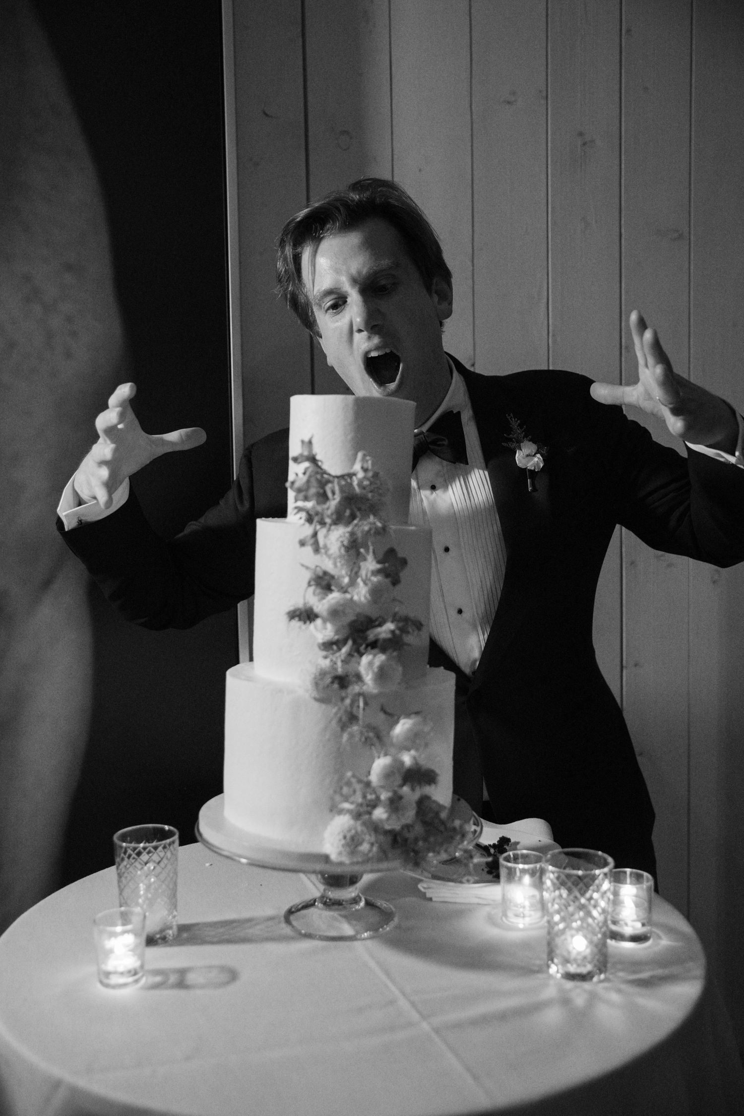 Excited groom posing dramatically with elegant white wedding cake decorated with cascading blue flowers.