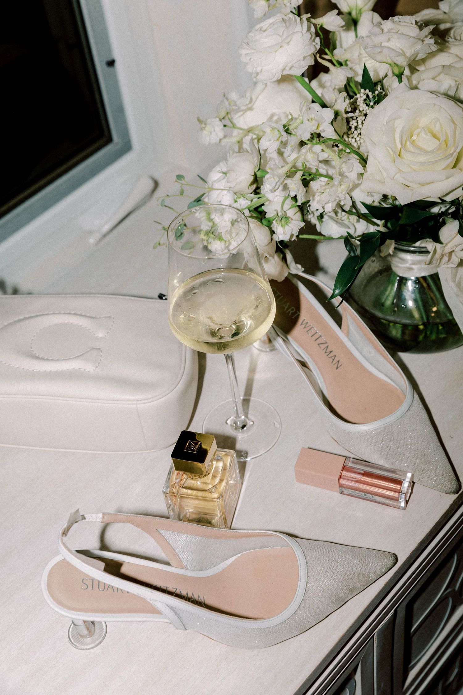 Wedding details featuring white roses, champagne, perfume, and elegant bridal shoes on a white surface.