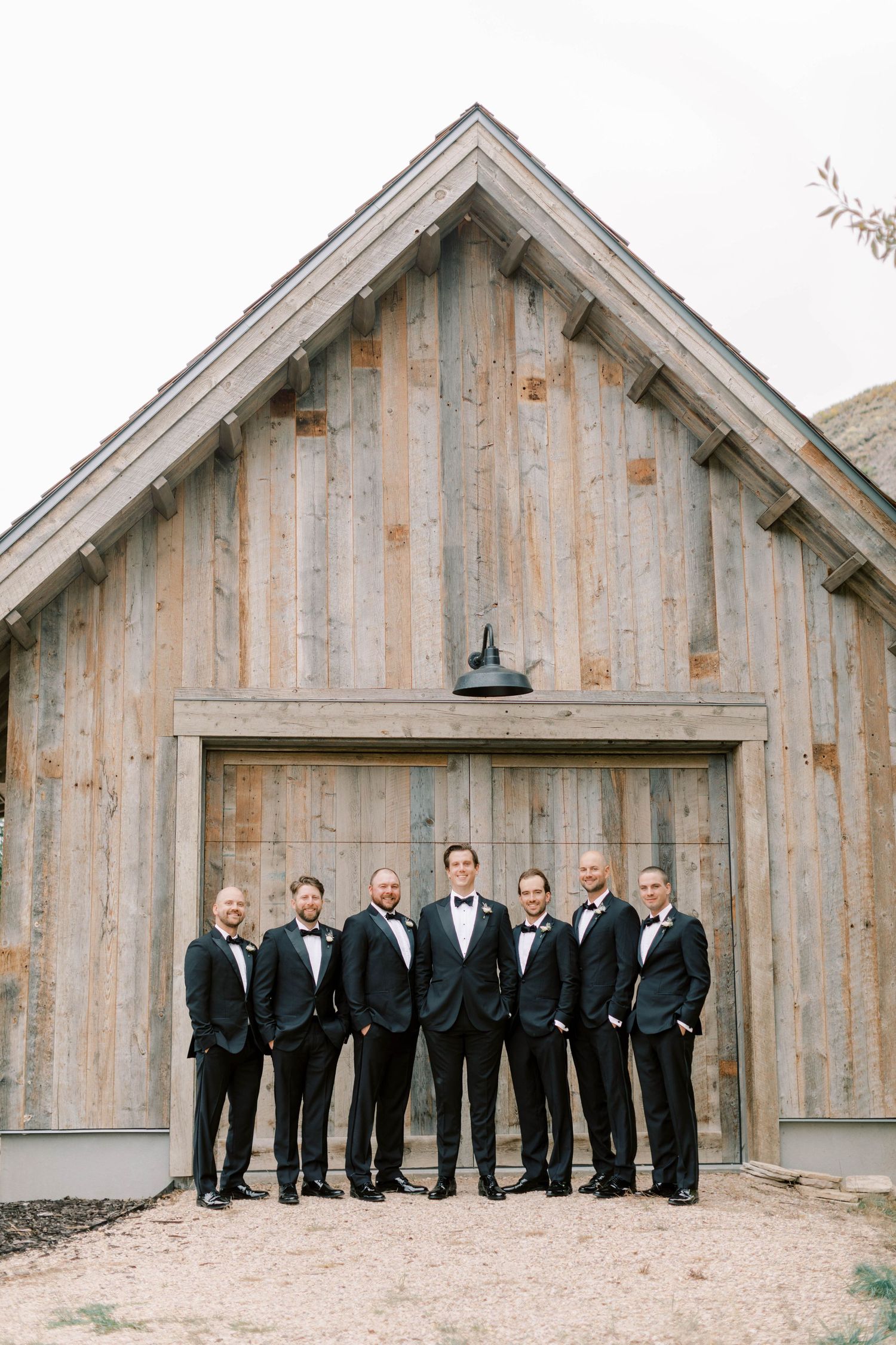 Groom and groomsmen in black tuxedos pose in front of rustic wooden barn door.