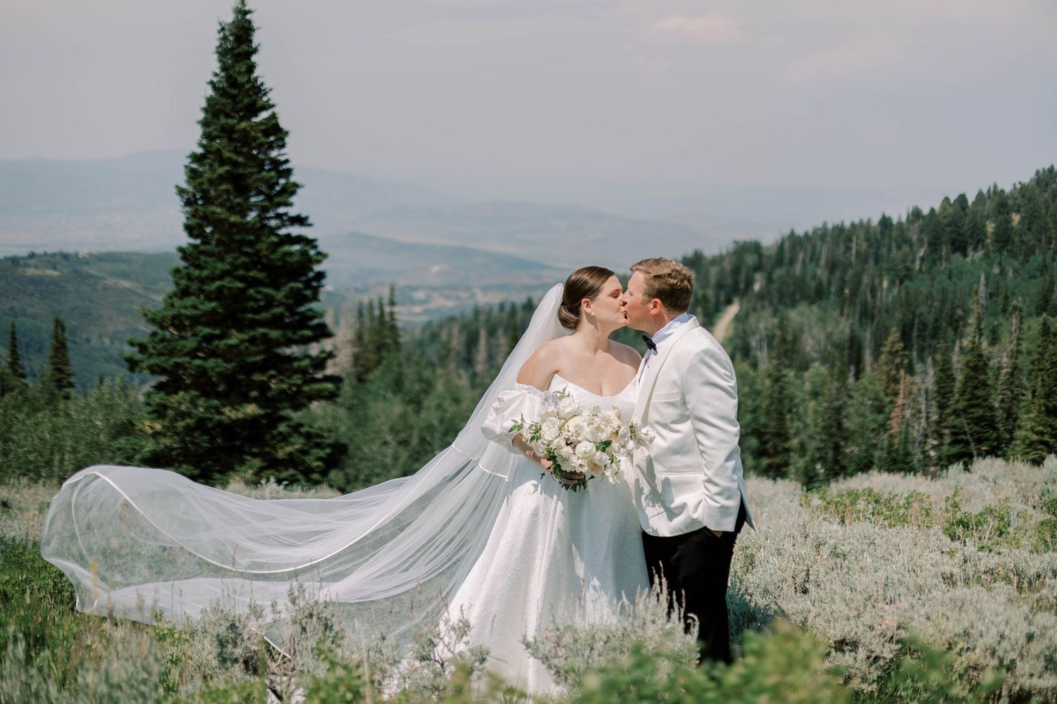 Romantic wedding portrait with flowing veil against mountainous backdrop and evergreen trees.