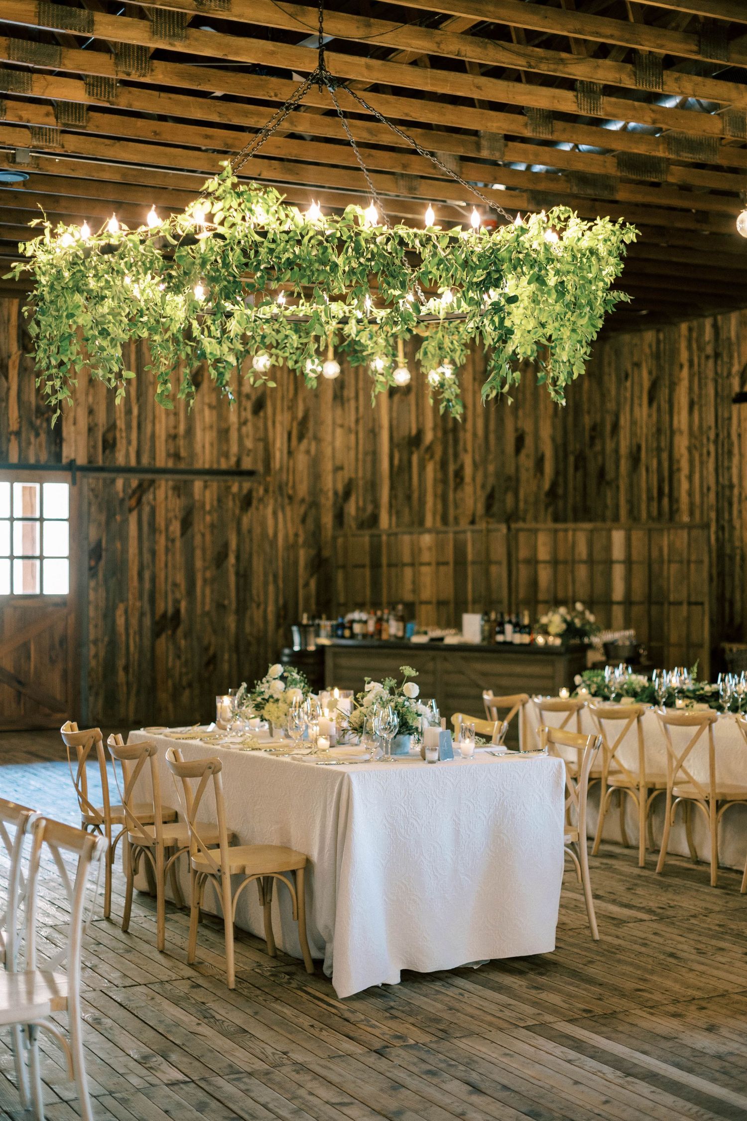 Rustic barn wedding reception with hanging greenery chandelier and white table settings.