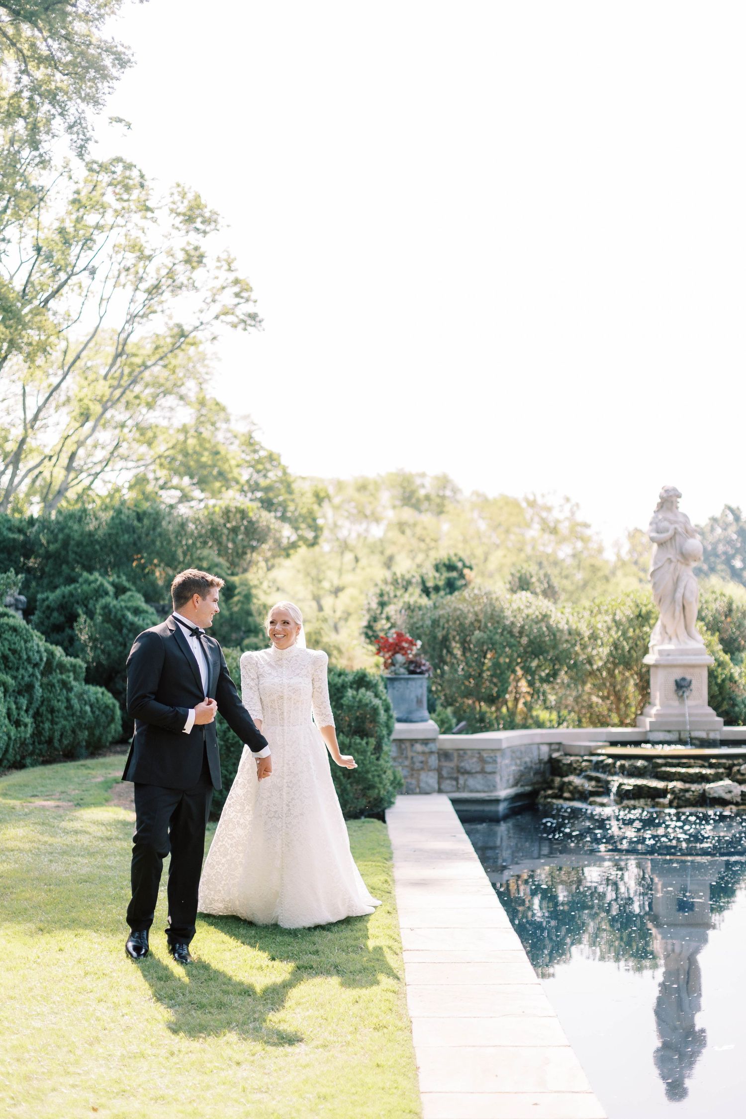Elegant wedding couple stands beside a reflective pool with classical stone statues in a manicured garden setting of Cheekwood Estate.
