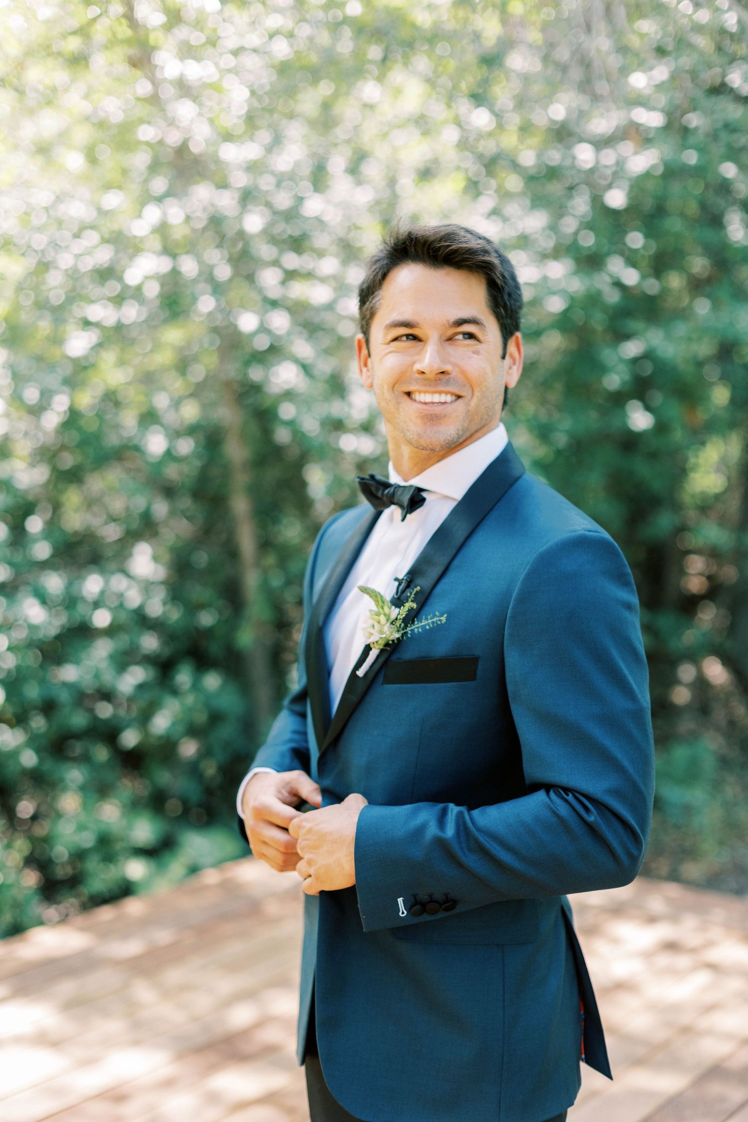 Groom in a navy blue tuxedo smiles outdoors against a blurred natural background.