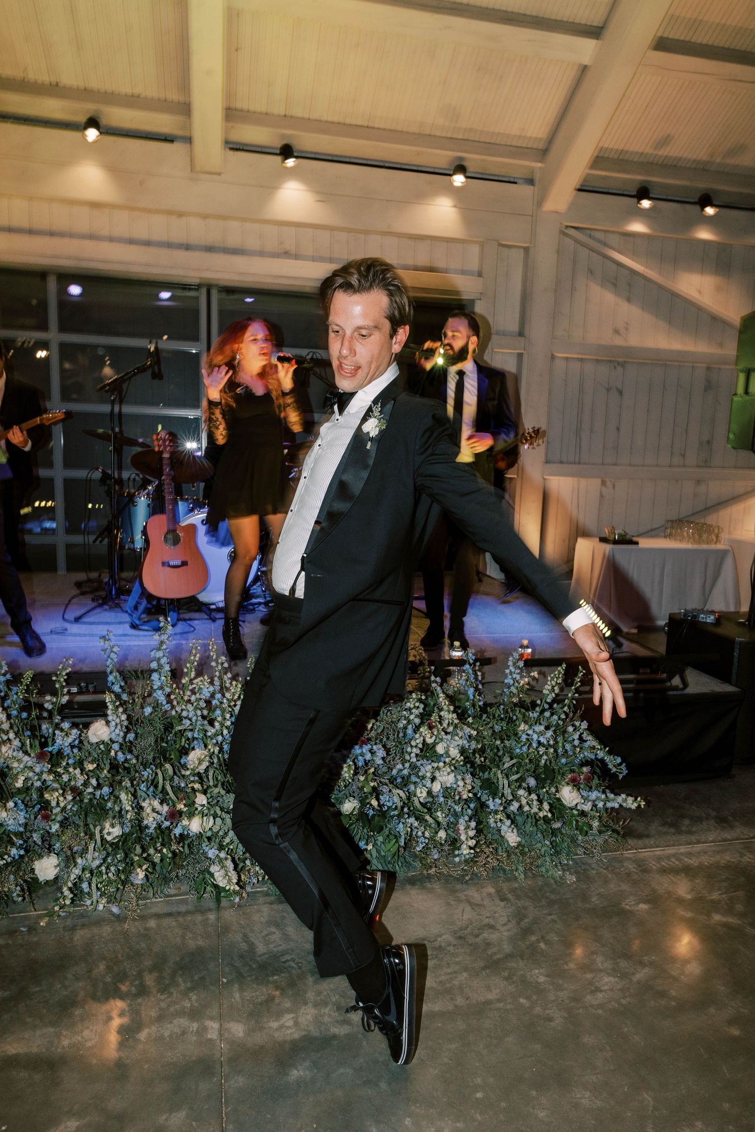A groom in a tuxedo jumps playfully on the dance floor with a live band setup in the background.