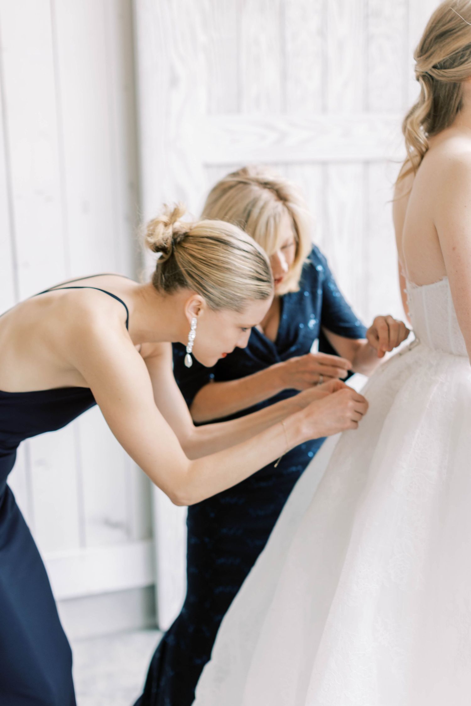 Bridesmaids in navy dresses help adjust the bride's wedding gown during preparation.