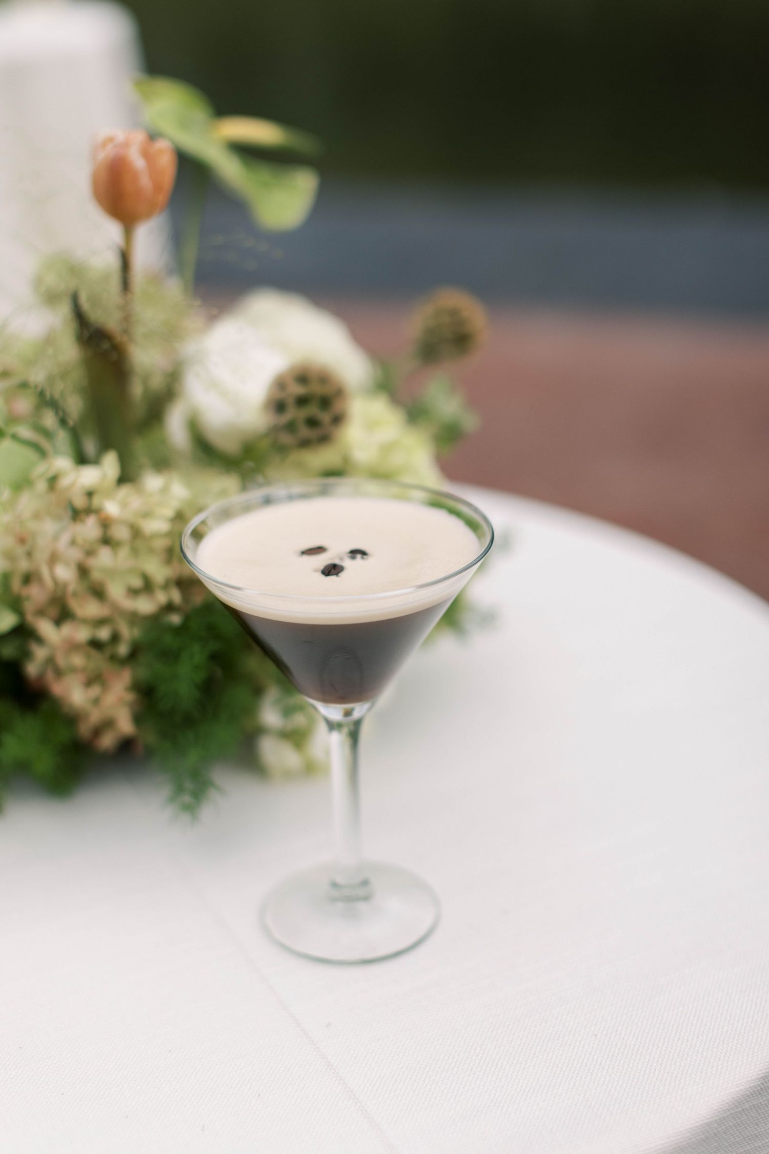 Elegant martini glass with creamy cocktail garnished with coffee beans beside floral arrangement.