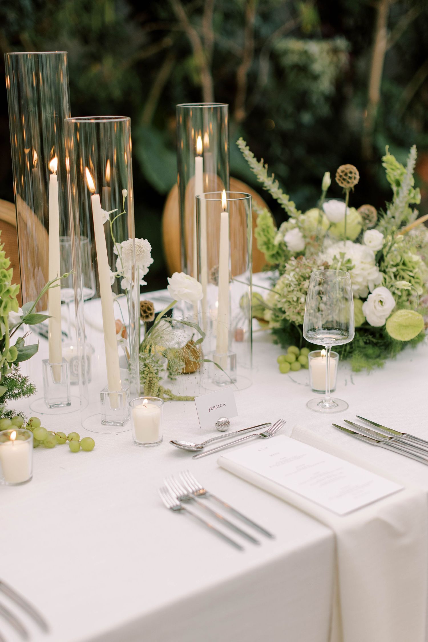 Elegant wedding table setting with tall glass candle holders, white flowers, and greenery against a dark background.