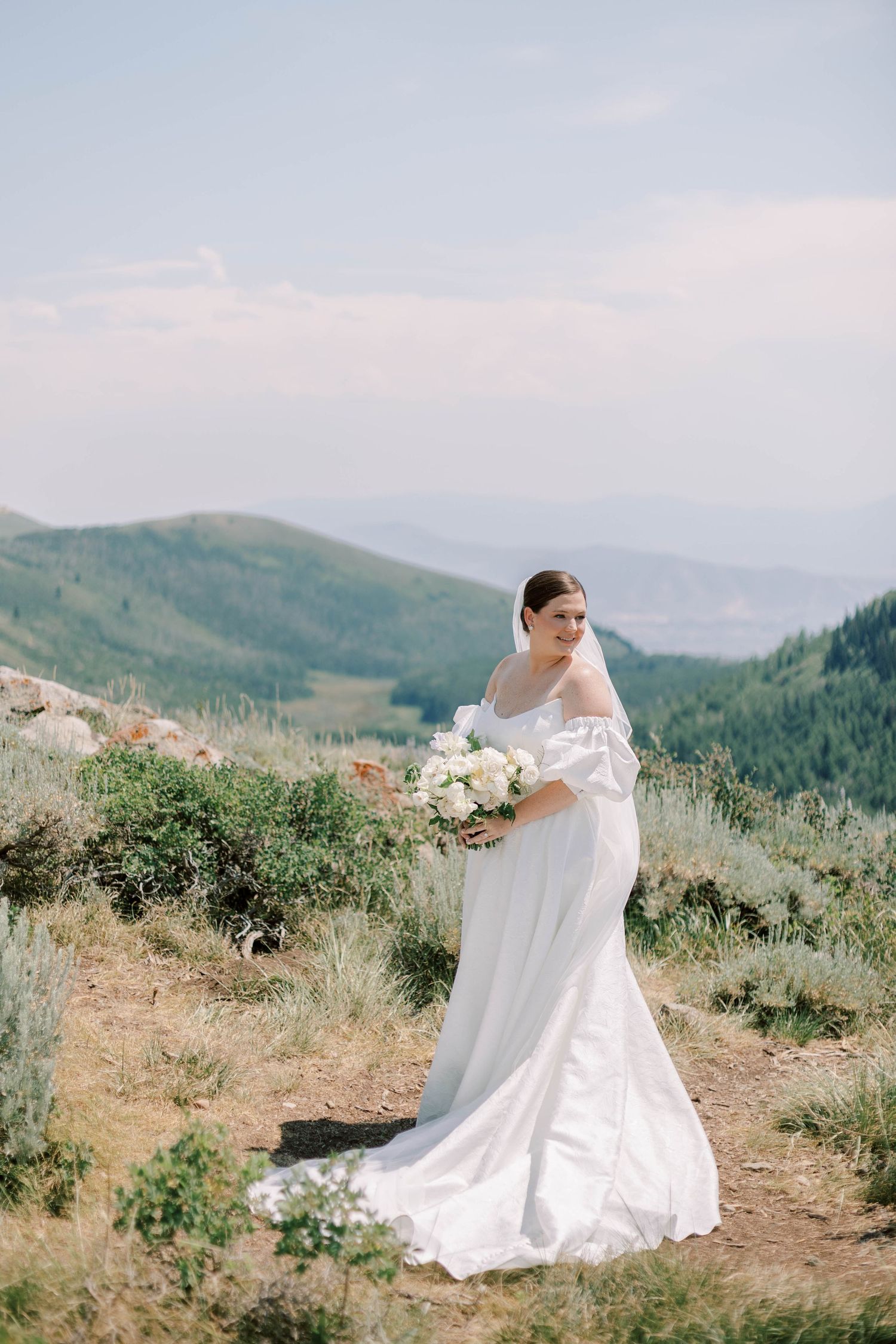 Bride in off-shoulder gown holds white bouquet on mountain trail.