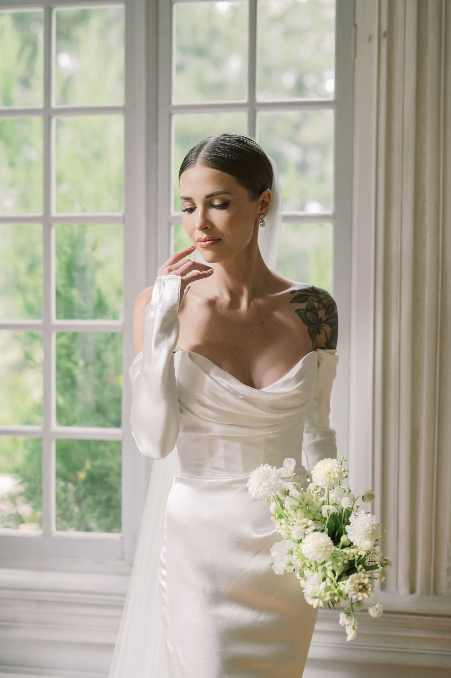 Elegant white wedding dress with off-shoulder design and delicate bouquet by sunny window.