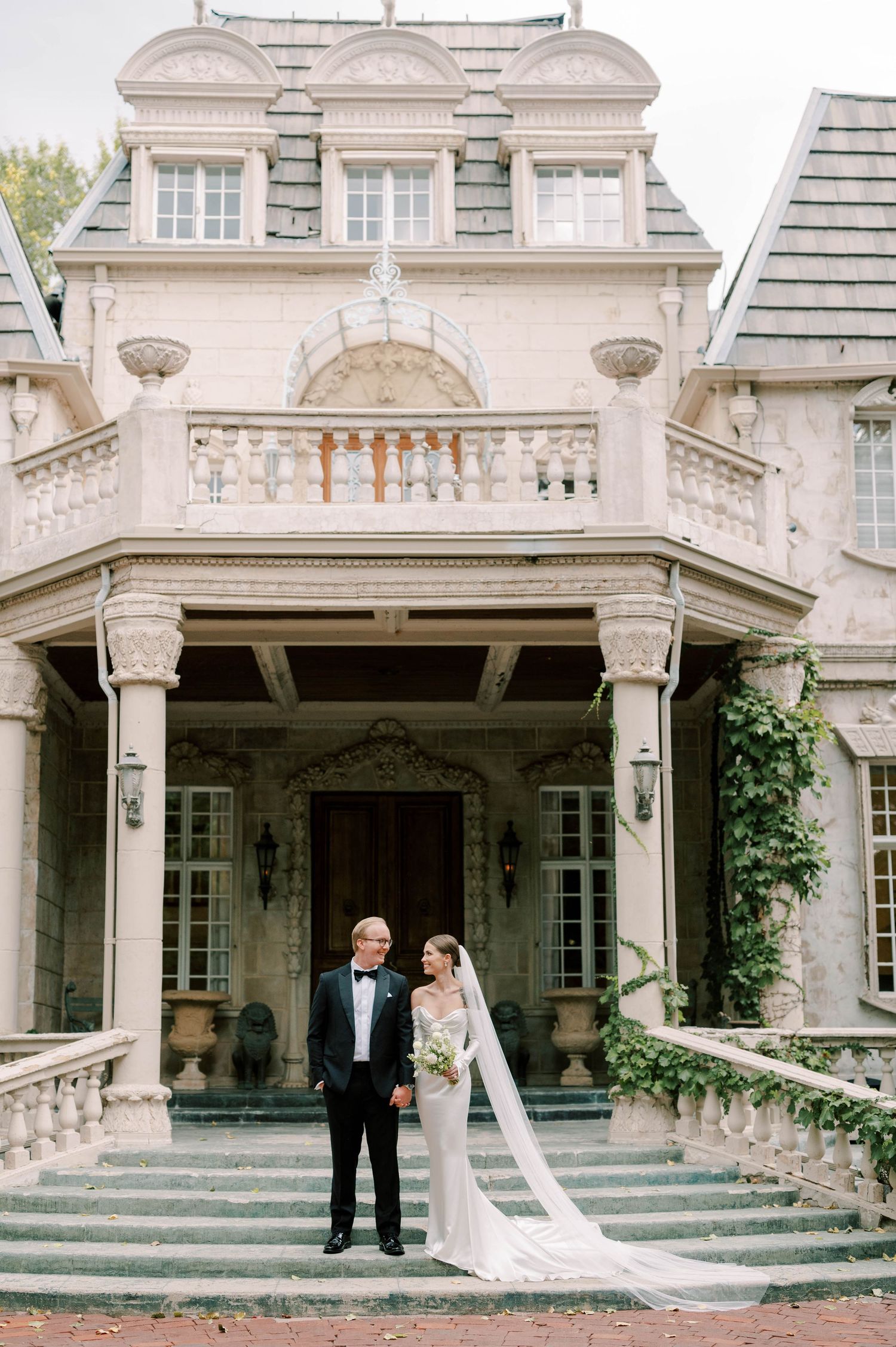 Elegant wedding portrait on steps of historic white mansion with columned entrance.