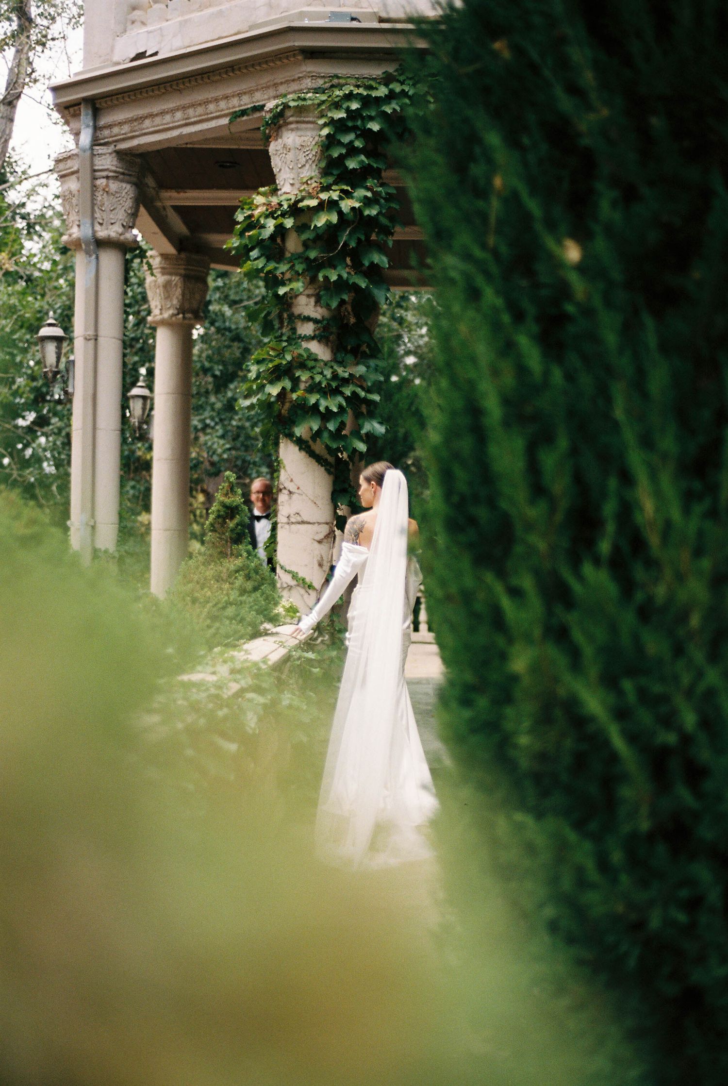 Bride in flowing white dress poses gracefully by stone pillars and cypress trees.