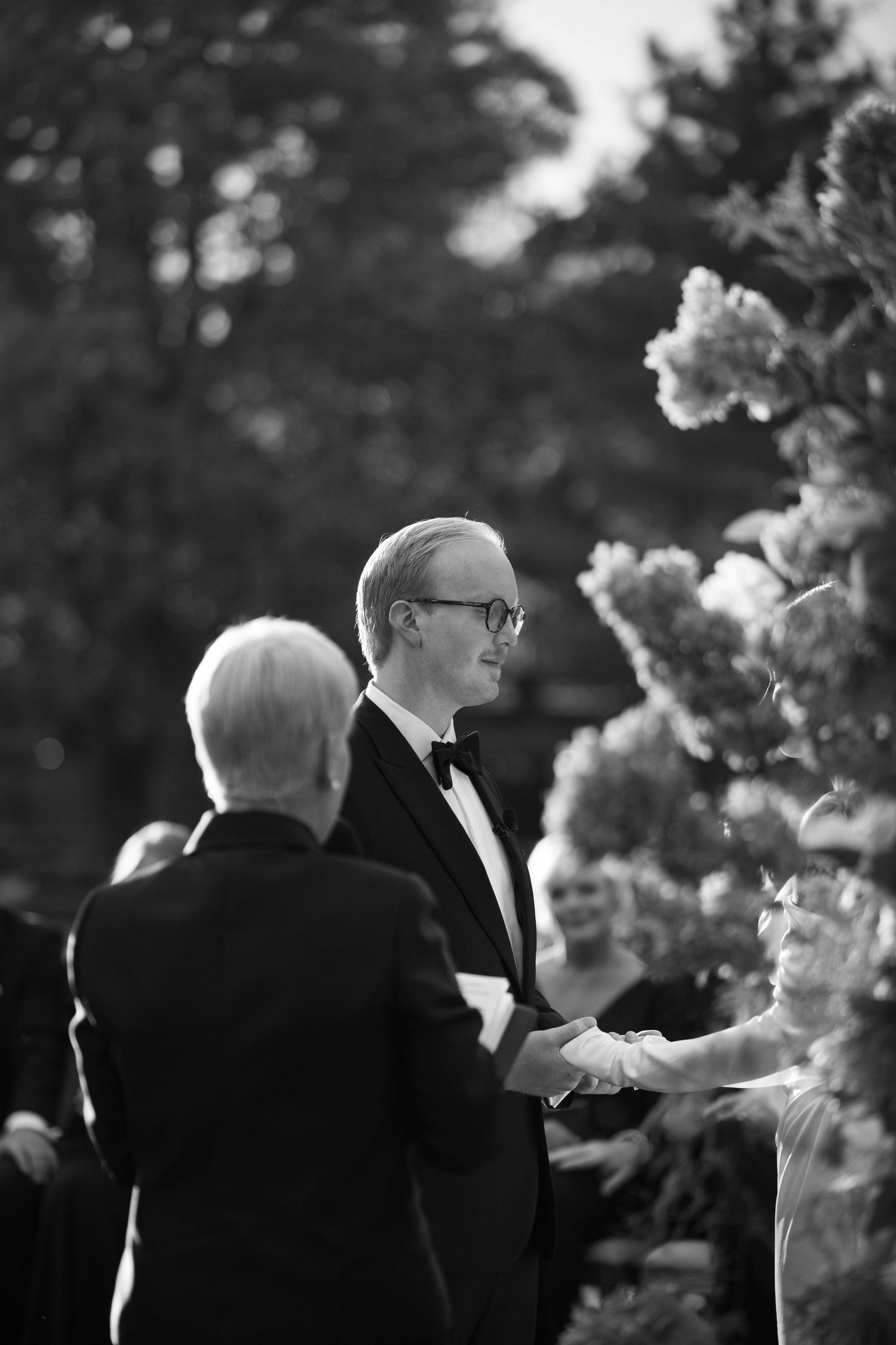Black and white photo of wedding ceremony moment with floral arrangements in soft focus.