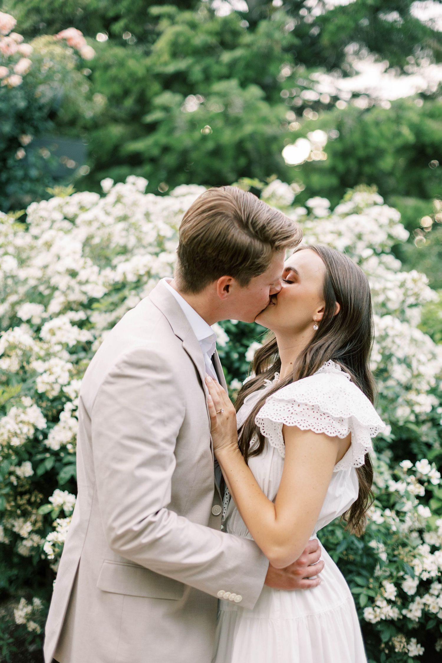 A couple shares a romantic kiss surrounded by blooming white flowers in a garden setting.