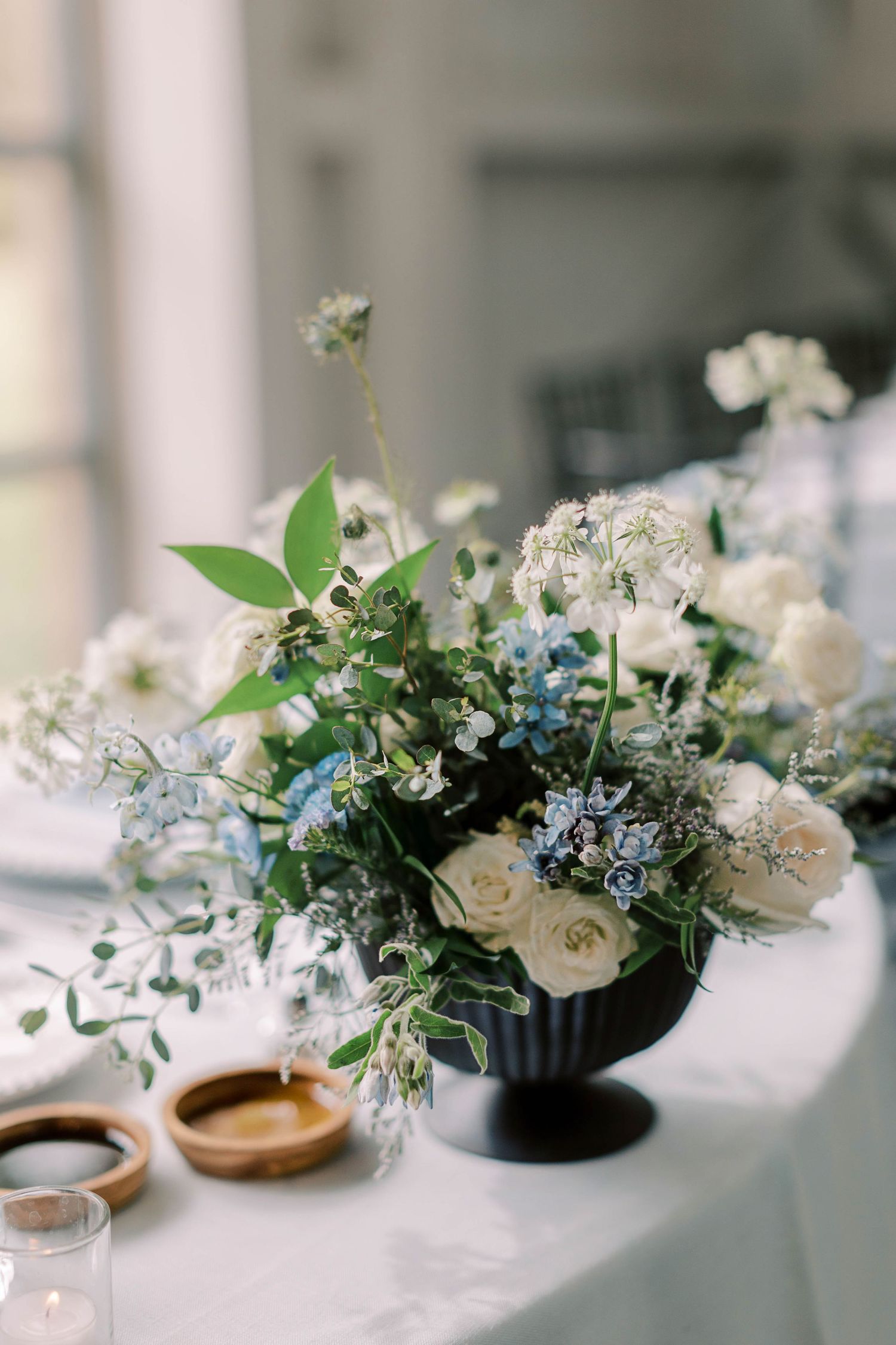 Elegant floral centerpiece with white roses and blue flowers in a black vase on a white tablecloth.