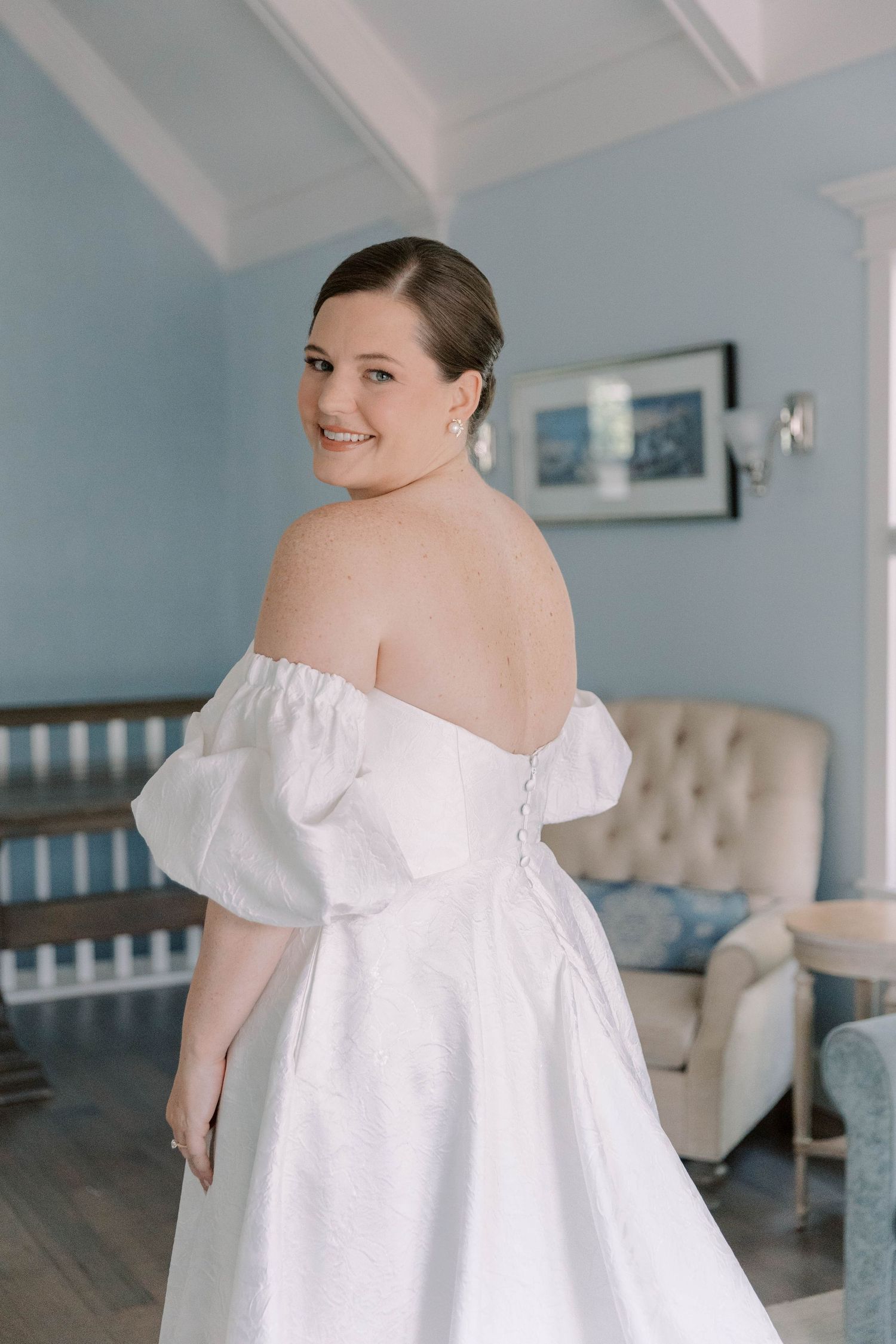 Smiling bride in an off-shoulder white wedding gown poses in a blue room.