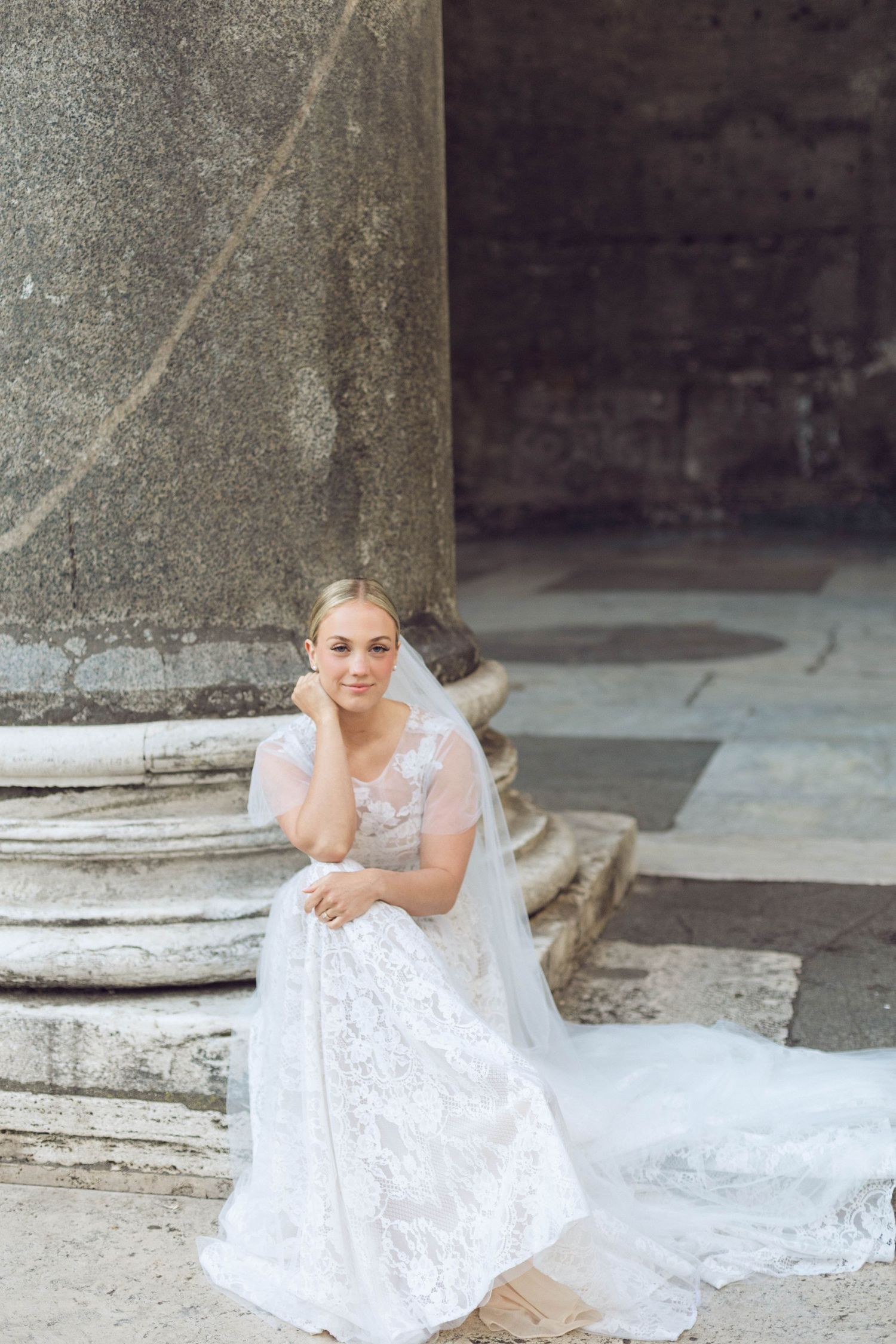 A bride in a lace white wedding dress and veil poses against classical stone columns.