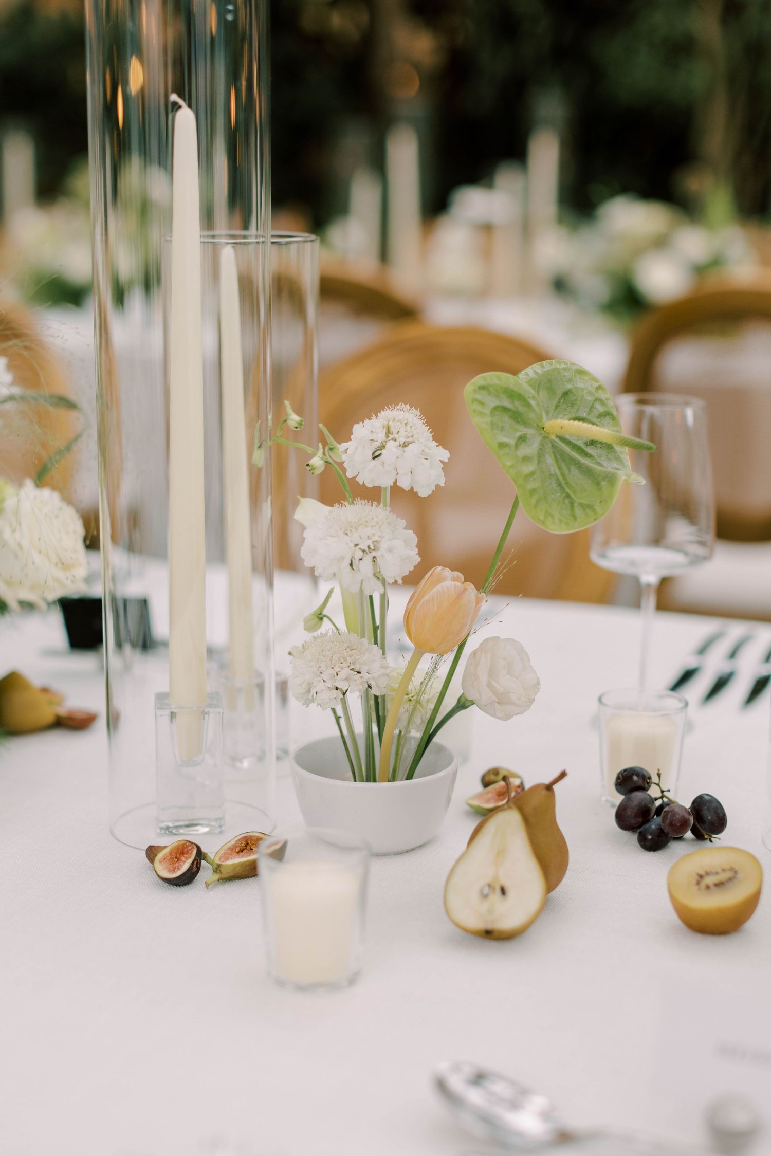 Minimalist table setting with white flowers, tall glass candle holders, and fresh fruit accents.