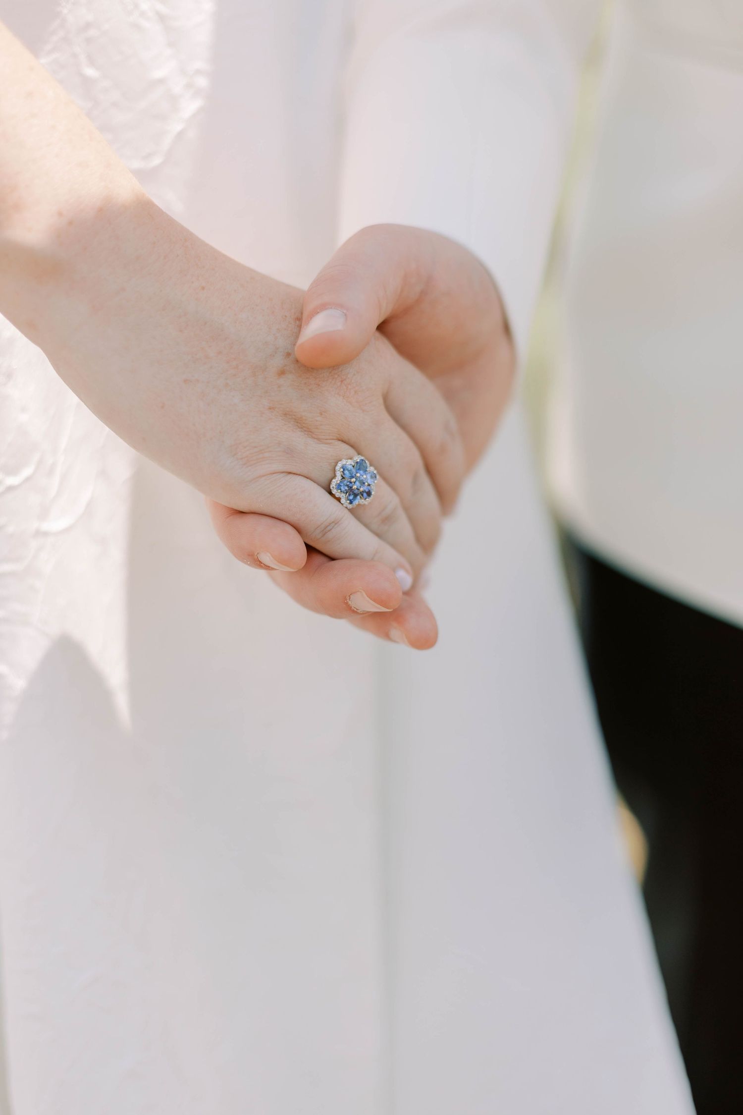 Close-up of clasped hands with a sparkling blue engagement ring against white fabric.