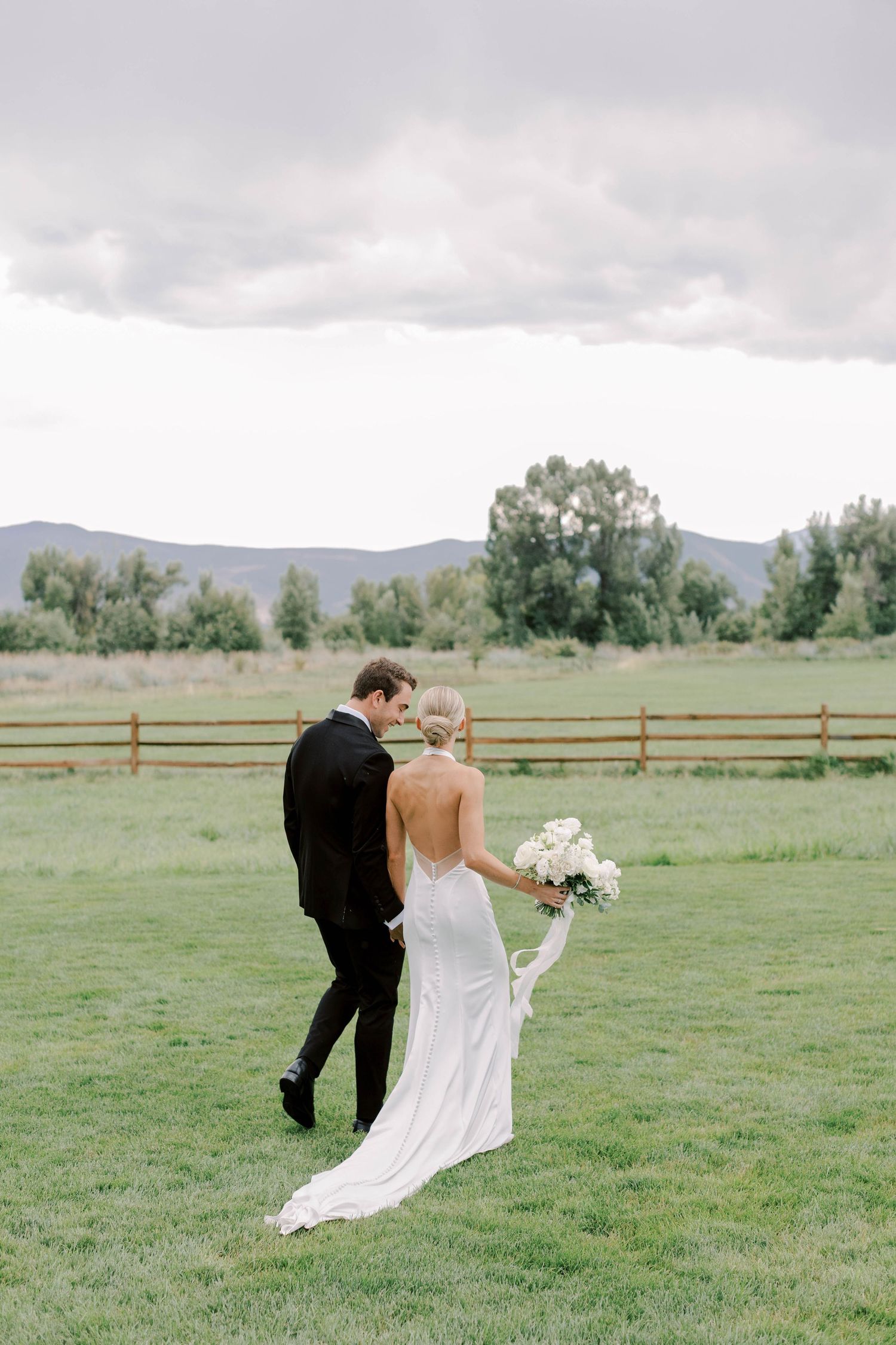 Newlyweds walk together on grass with mountain backdrop and white bouquet.