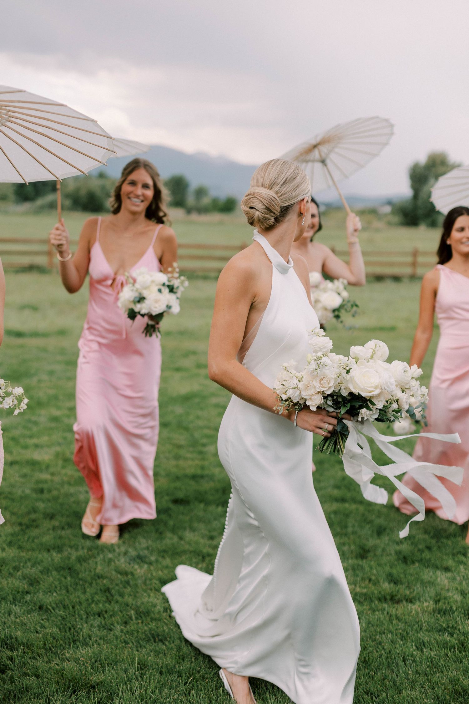 Wedding party members with white parasols and bouquets walking on grass with mountains behind.