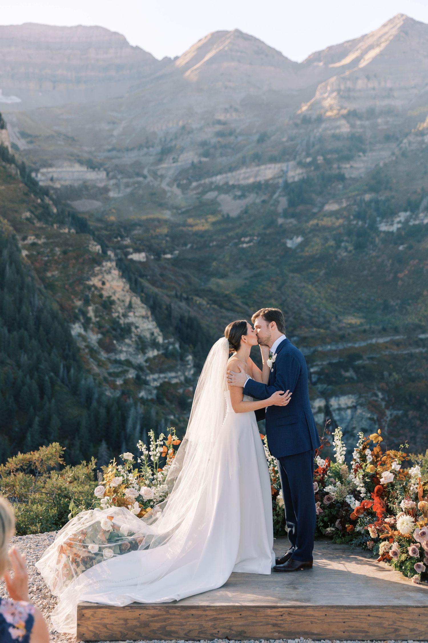 A couple embraces during their mountain wedding ceremony surrounded by colorful flowers.