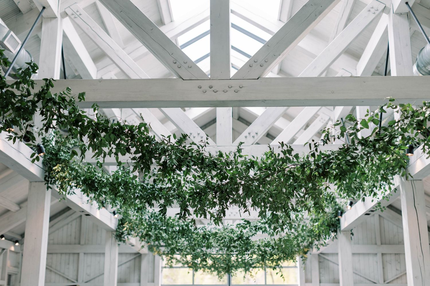 White barn wedding venue interior with elegant greenery garlands hanging from exposed beam ceiling.