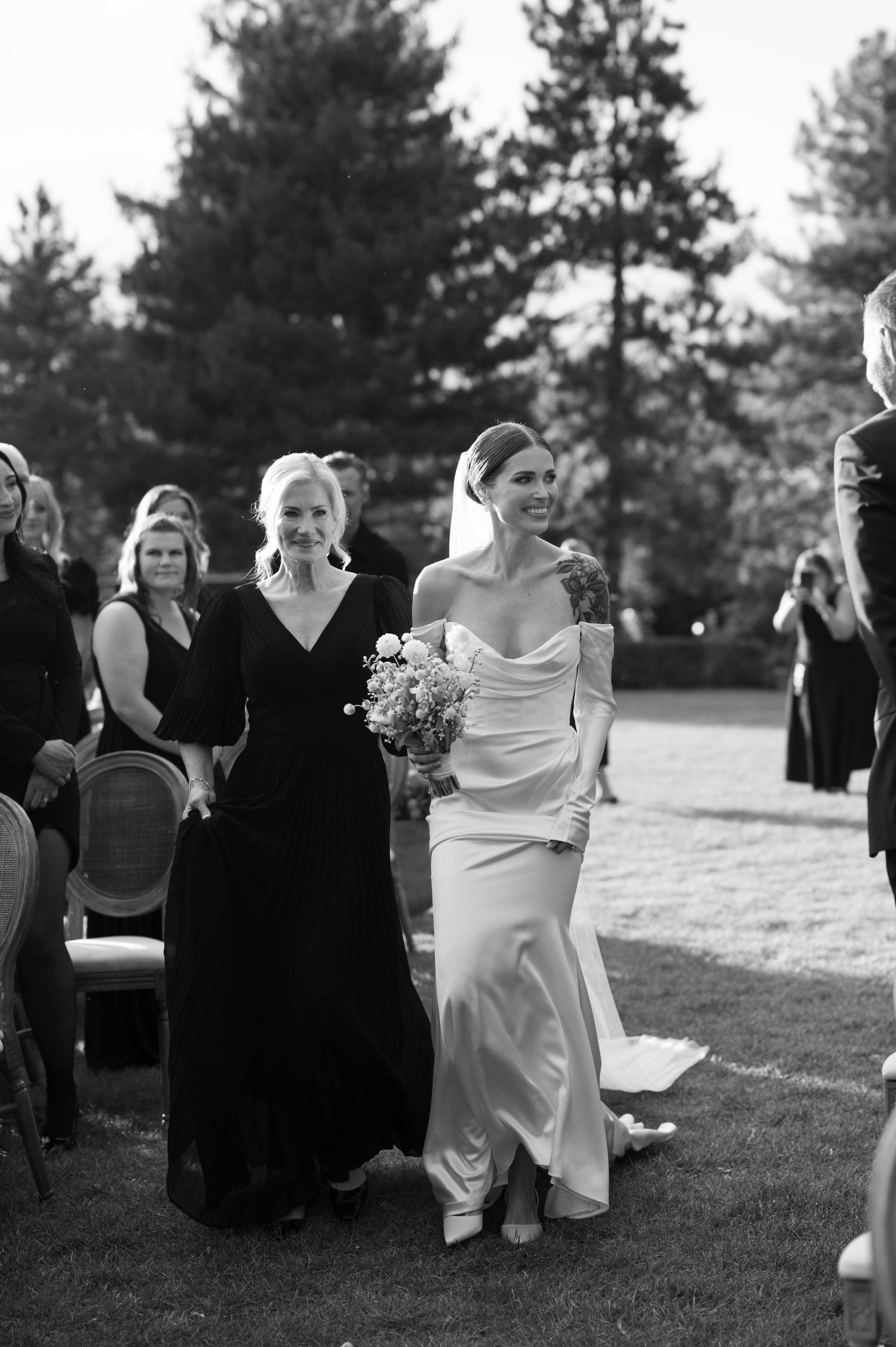 Wedding party in black dresses gathers around a bride in an elegant white gown against a backdrop of evergreen trees.