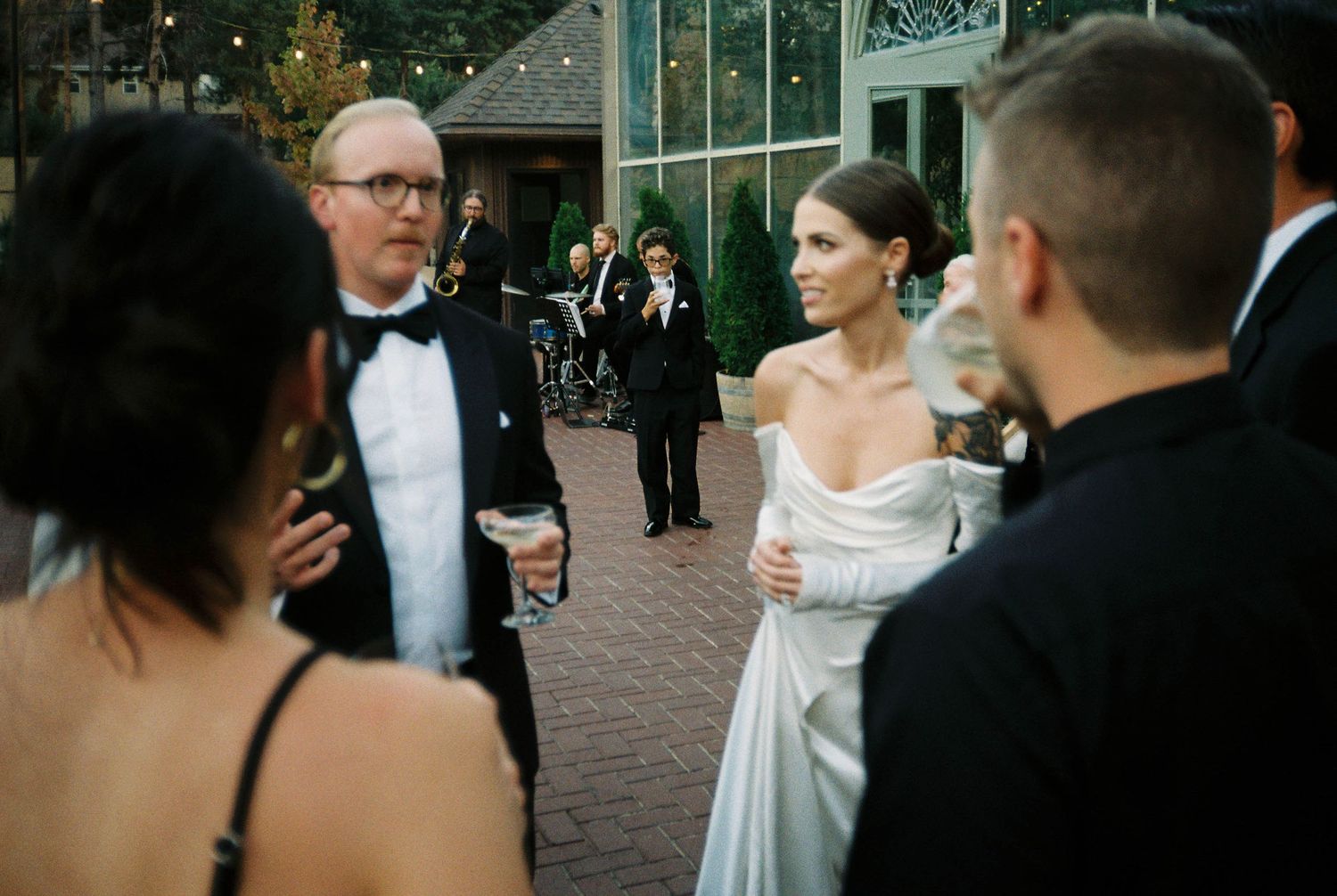 Elegant wedding guests in formal attire gather outside a glass conservatory during an evening reception.